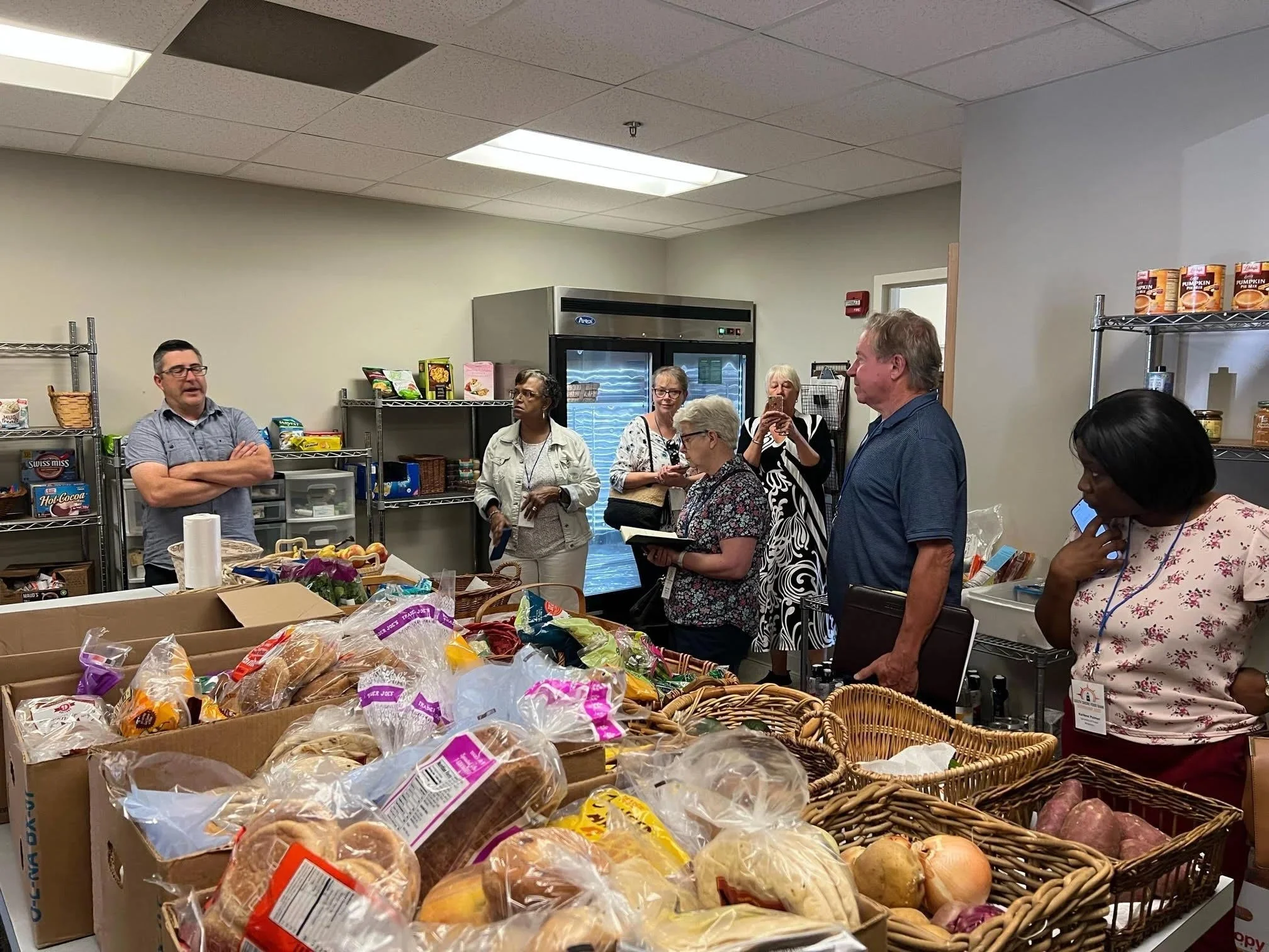 A group of people gathered in a food pantry, standing around a table filled with bread, baked goods, and vegetables. Some are listening to a speaker, while others are taking notes or photos.