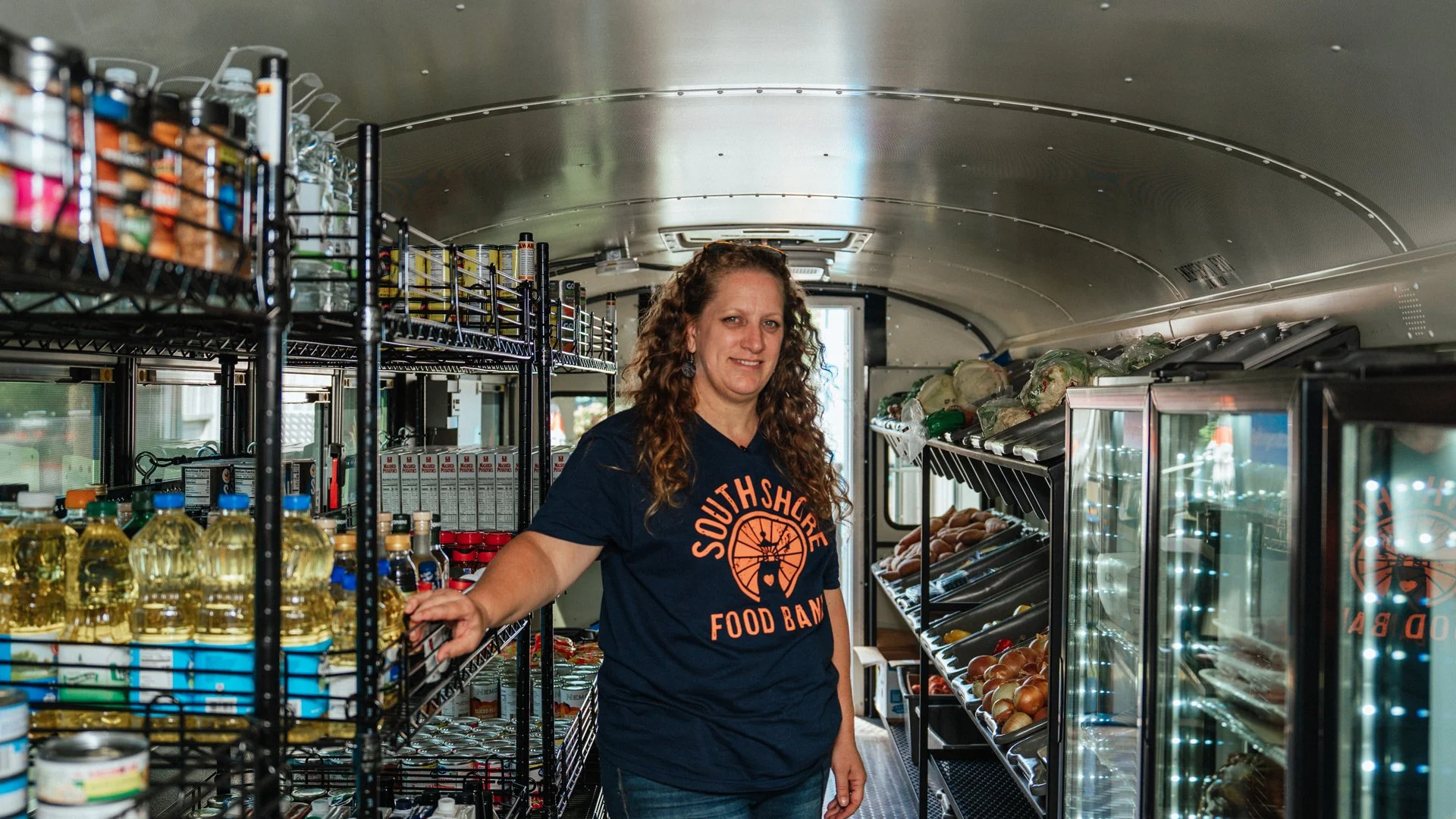 A woman with curly hair wearing a navy blue T-shirt with 'South Shore Food Bank' logo standing inside a mobile food pantry with shelves stocked with canned goods, bottles of oil, vegetables, and other food items.