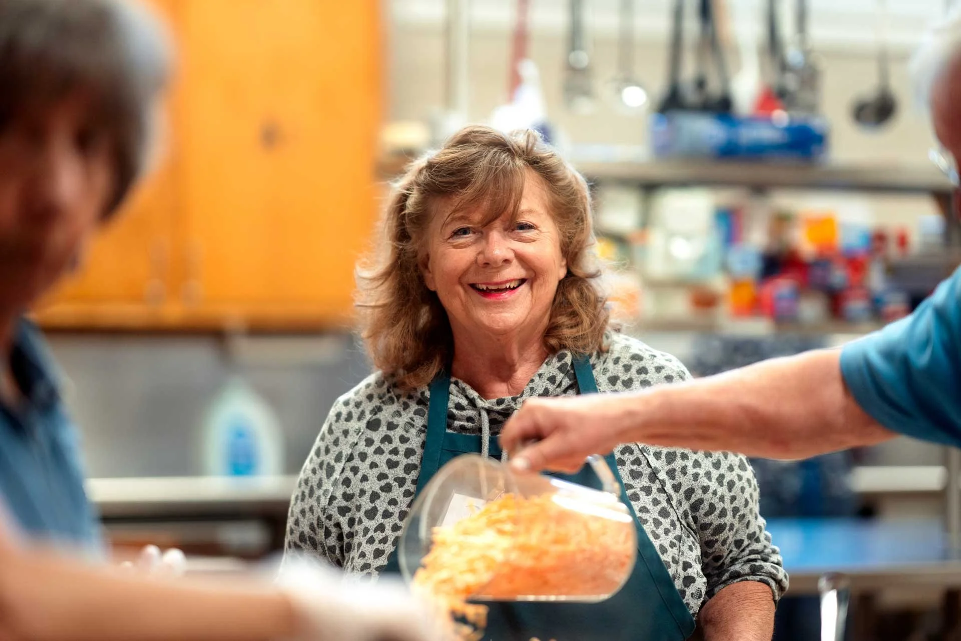A smiling woman in a kitchen, wearing a teal apron and a patterned shirt, receiving food from another person. The background shows kitchen shelves and utensils.