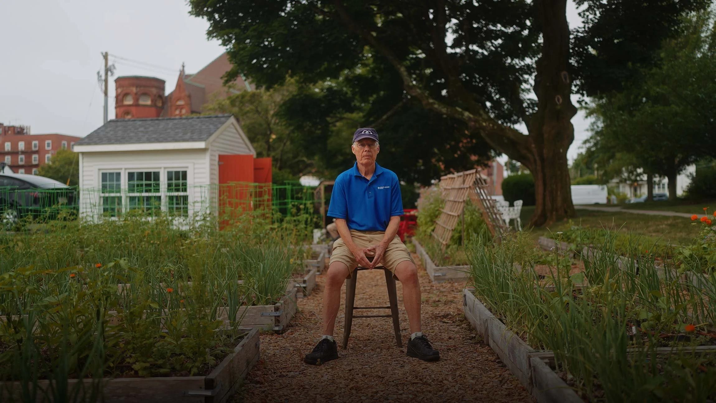 An elderly man sitting on a stool in a community garden with raised beds, trees, and a white shed in the background.