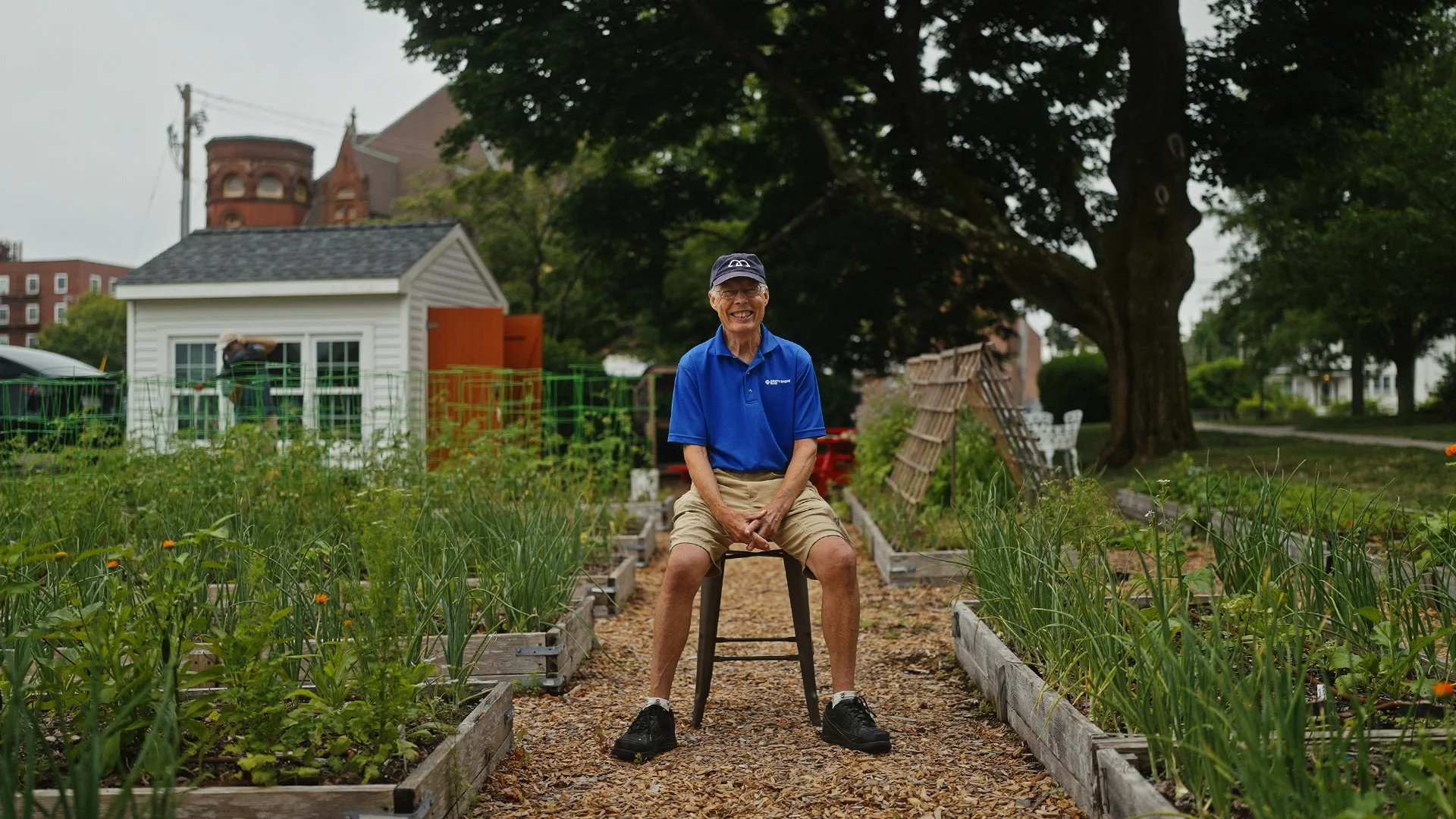 An man wearing a blue polo shirt, khaki shorts, and black sneakers sitting on a stool in a garden with raised vegetable beds and green trees in the Weymouth Pantry Garden.