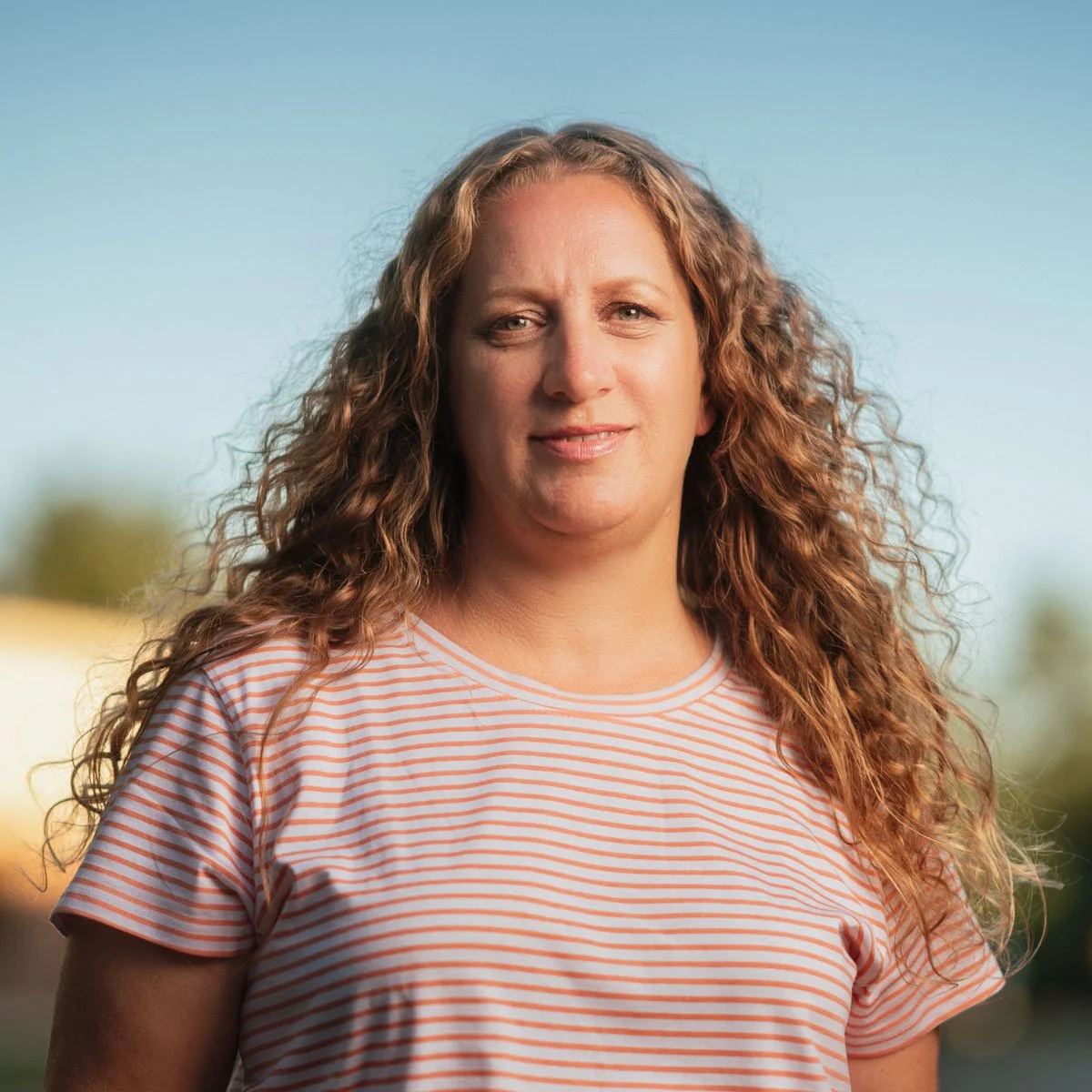 A woman with long curly hair wearing a white and orange striped shirt outdoors with a blue sky and blurred background.