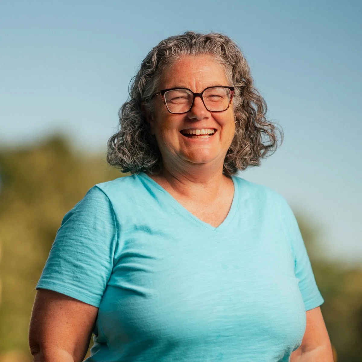 A smiling middle-aged woman with curly gray hair, glasses, and a light blue t-shirt outdoors with a blurred background of trees.