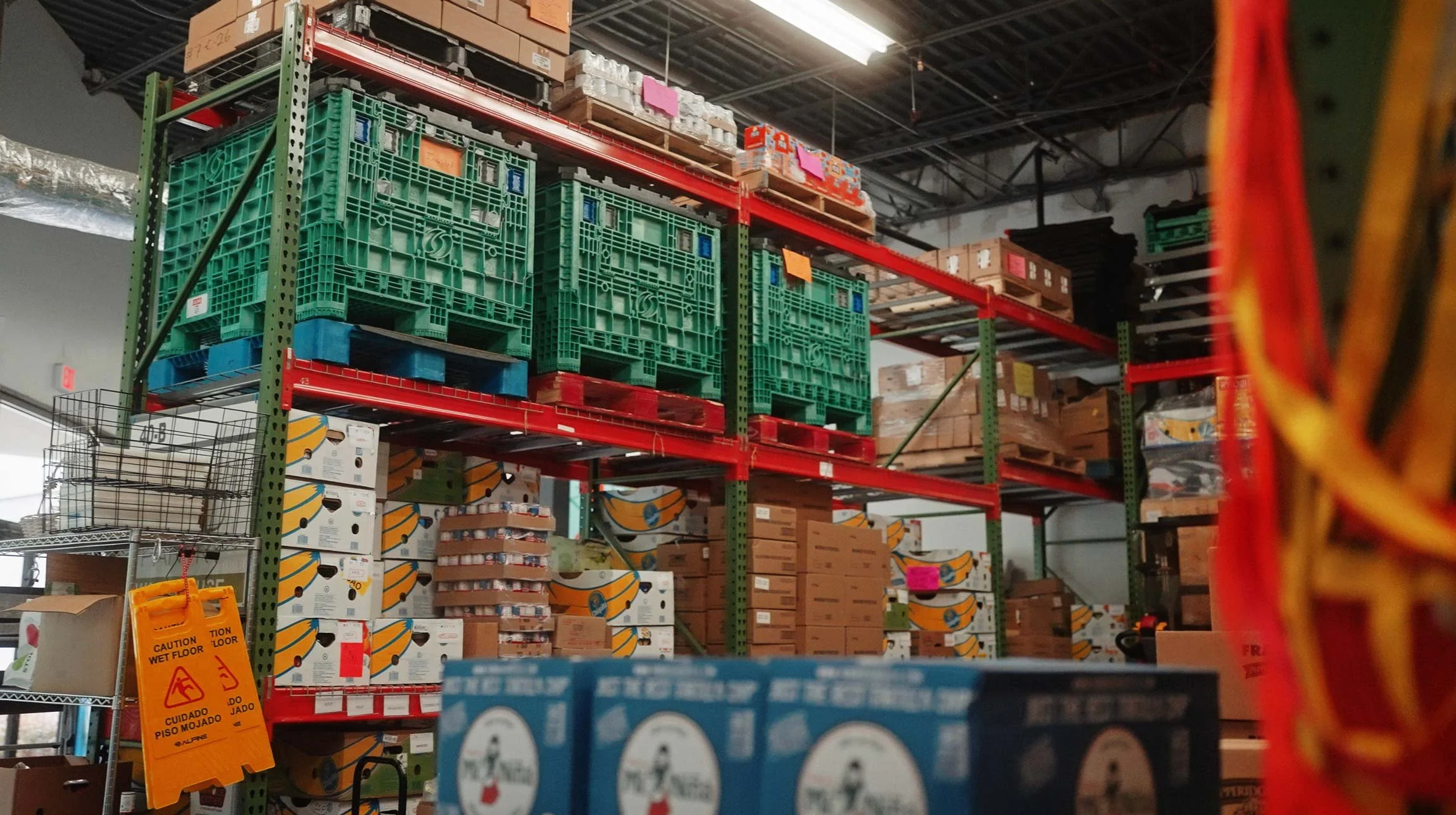 Warehouse aisle with shelves filled with boxes, crates, and pallets. Green crates on top shelves, and presence of a yellow caution sign on the floor. Various boxes and packages stacked on metal shelves and on the floor.