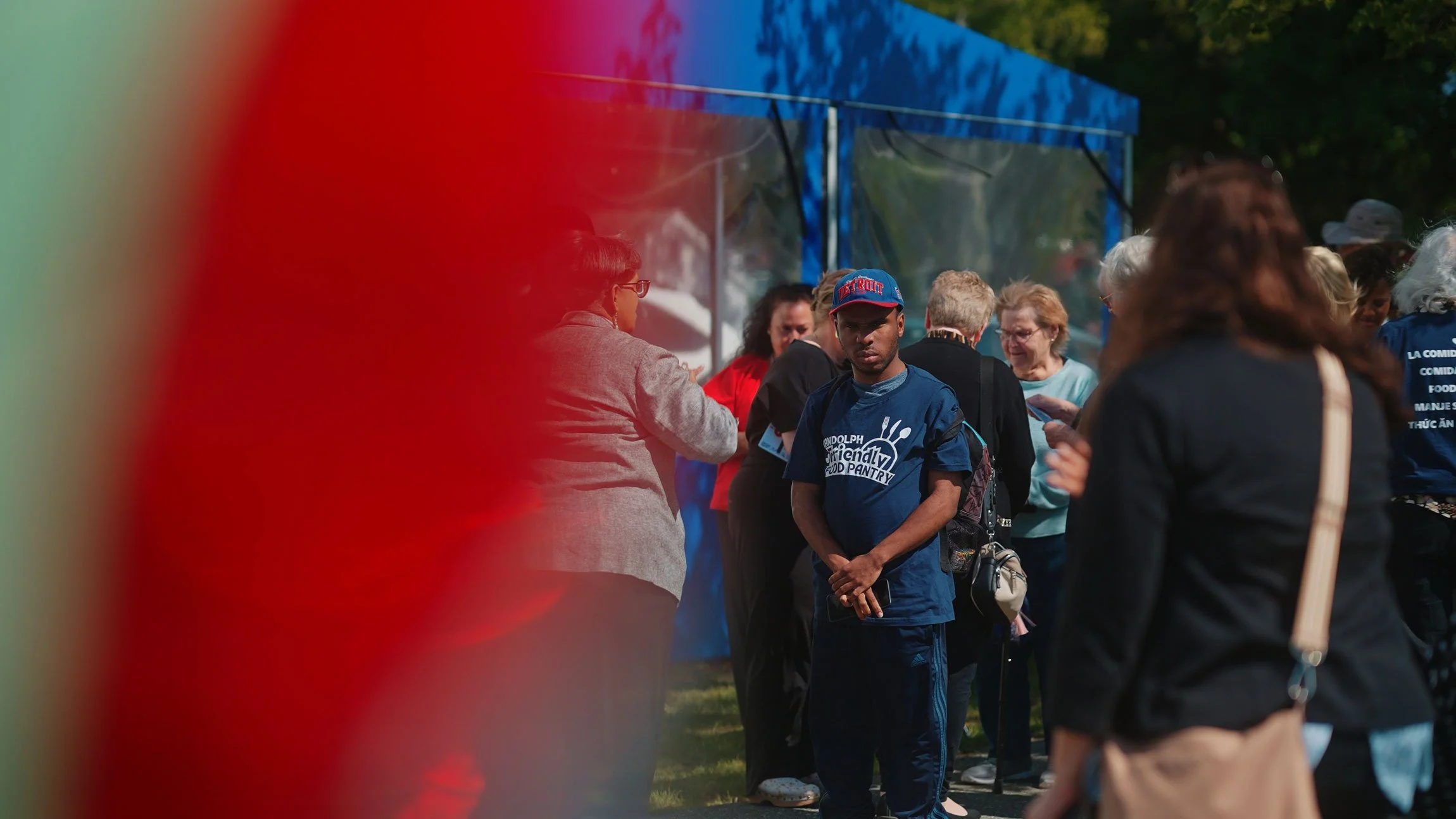A group of diverse people waiting in line outdoors near a blue tent on a sunny day.