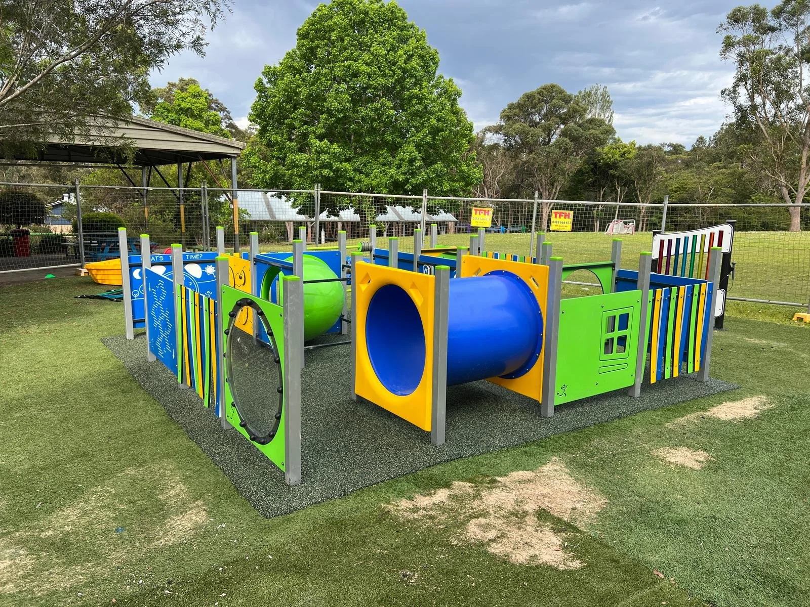 Colorful modern playground area with tunnels, climbing structures, and fencing on a grassy field, surrounded by trees and a cloudy sky.