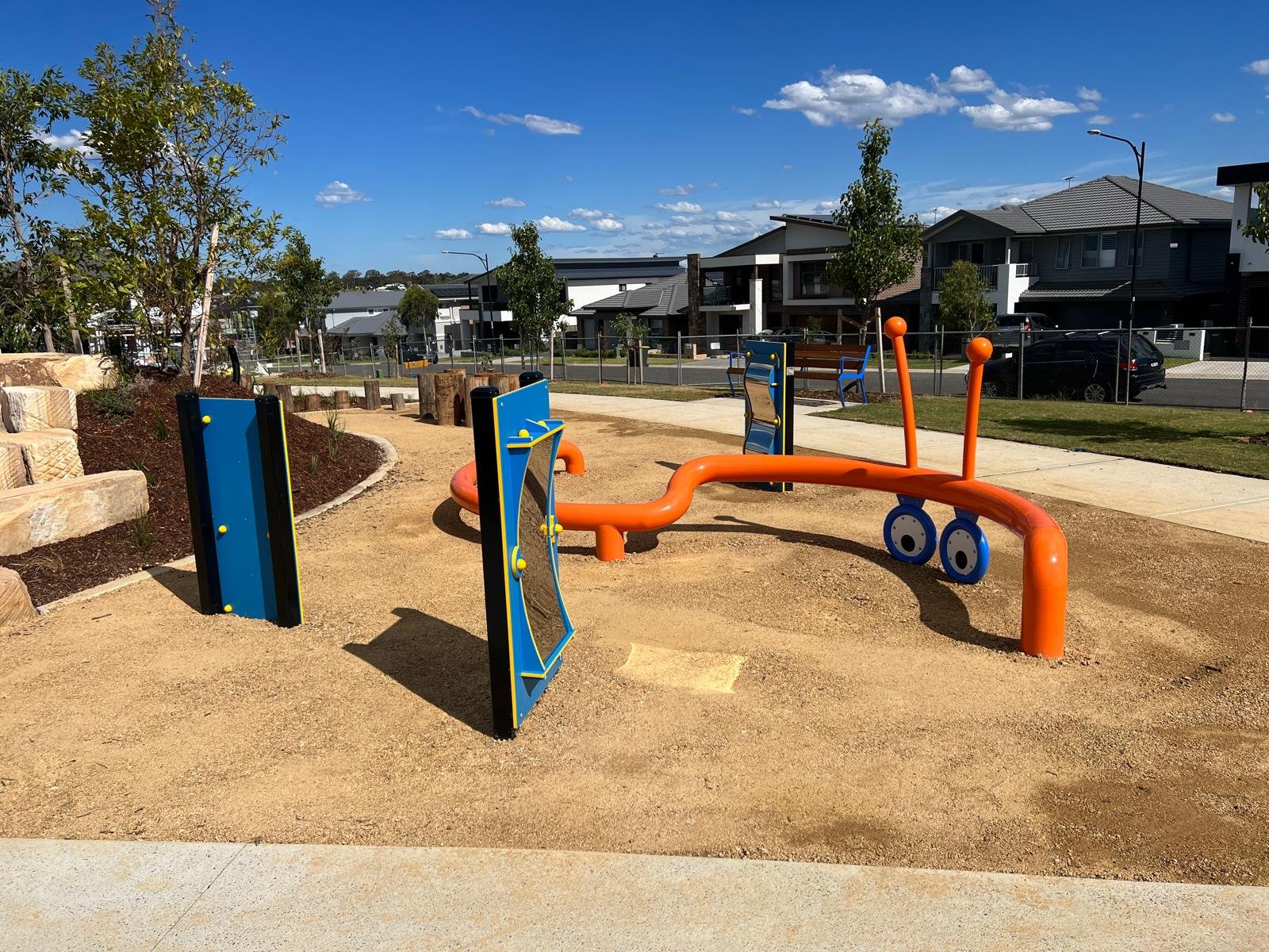 Children's playground with orange seesaw, climbing wall, and blue steering wheel at a park in a residential neighborhood under a clear blue sky.