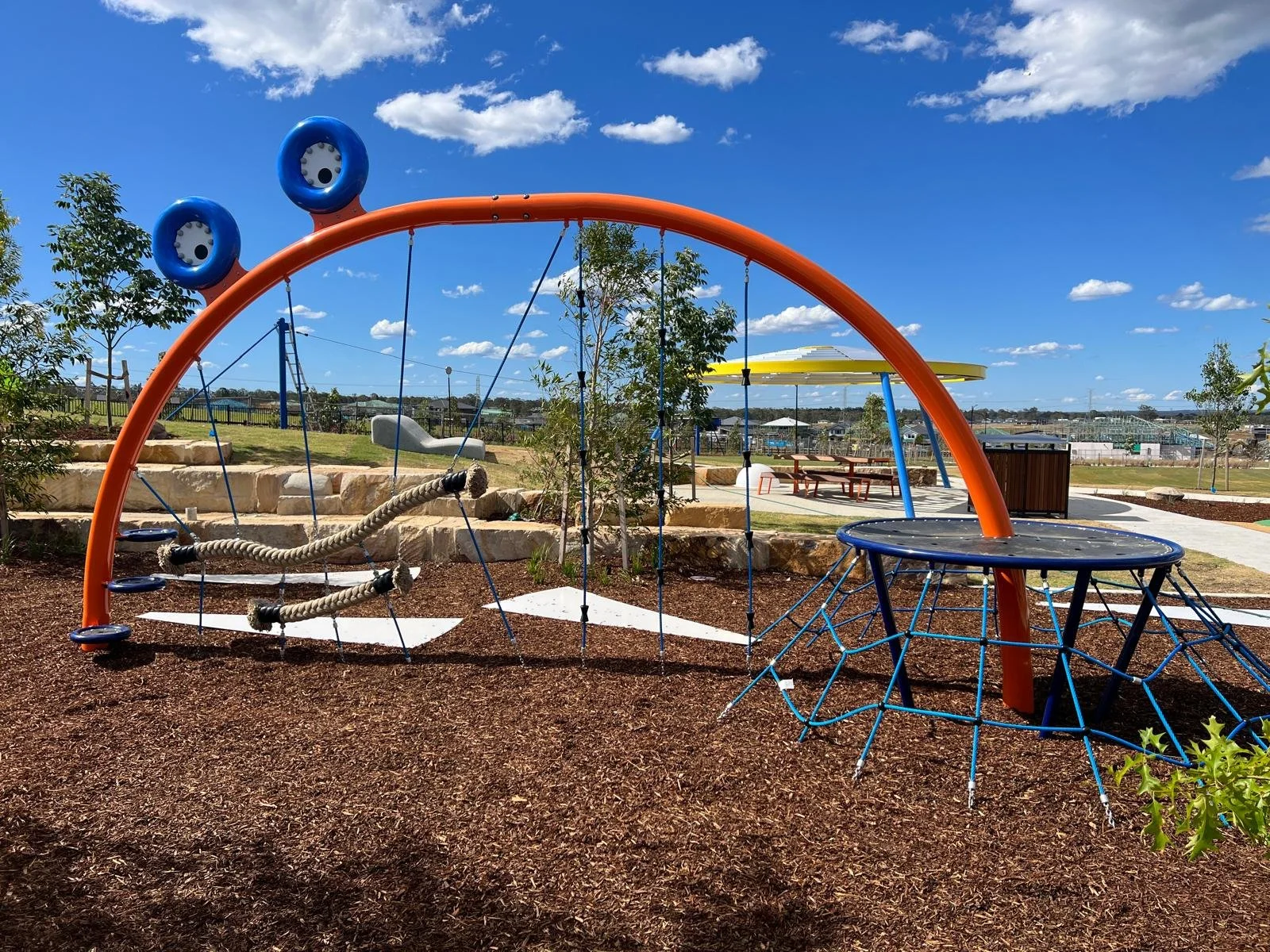 Colorful playground equipment with climbing ropes and a rubber surfacing under a blue sky with clouds.