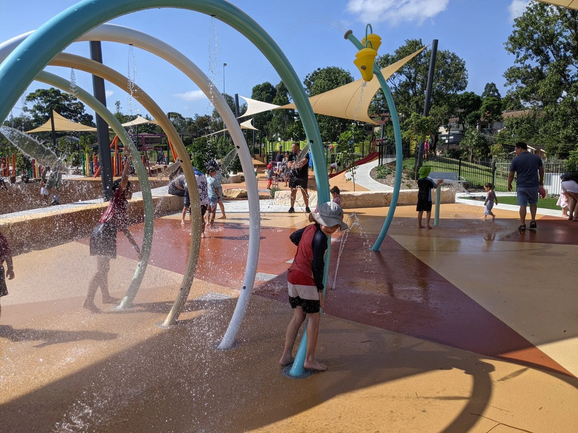 Children playing at a water park with spray fountains, colorful structures, and shade sails in a sunny outdoor setting.