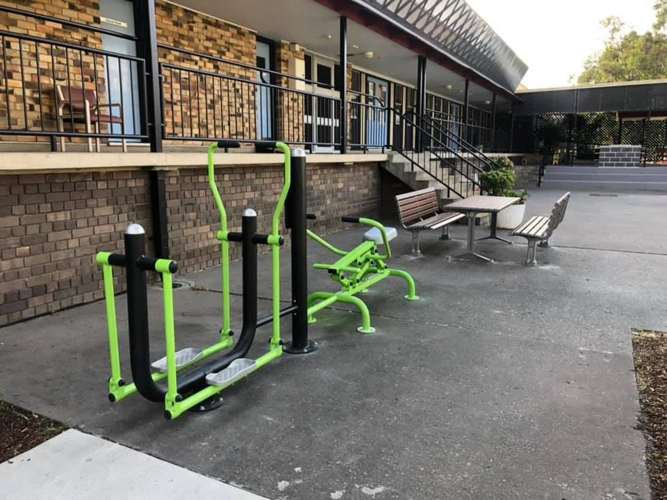 Outdoor exercise equipment with green and black components, two benches with seating and a table, against a brick building with large windows and stairs, near a landscaped area.