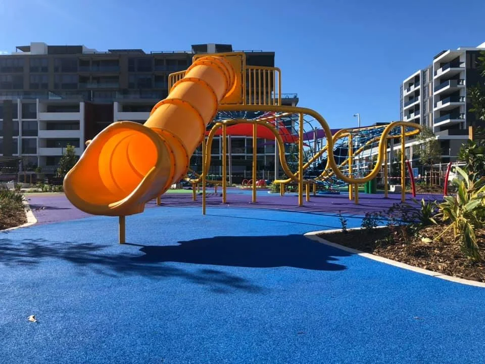 Colorful playground with a yellow slide, climbing structures, and a blue rubberized safety surface, surrounded by modern high-rise apartment buildings under a clear blue sky.