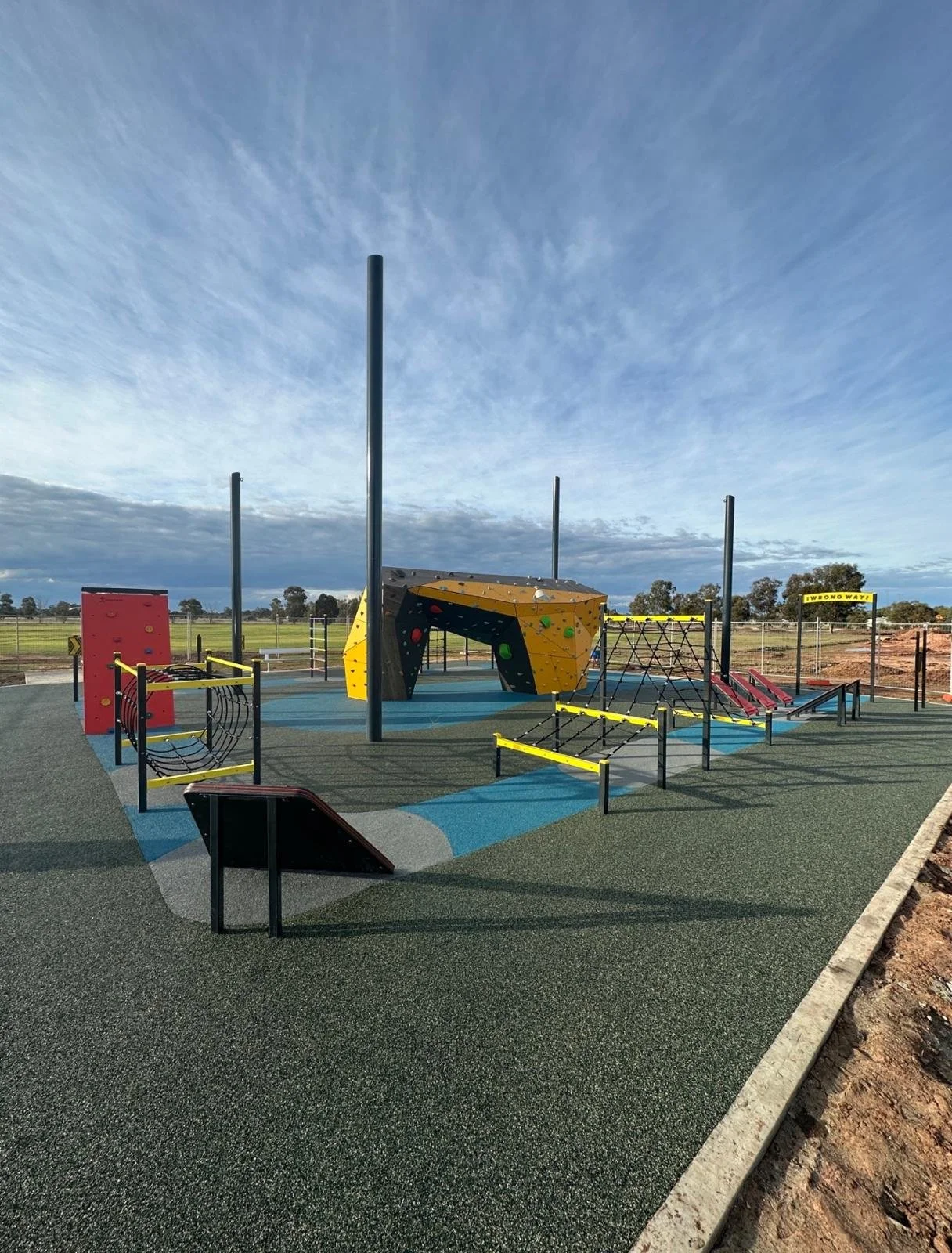 Empty outdoor playground with climbing structures, horizontal bars, and a blue and green rubberized surface, under a cloudy sky.