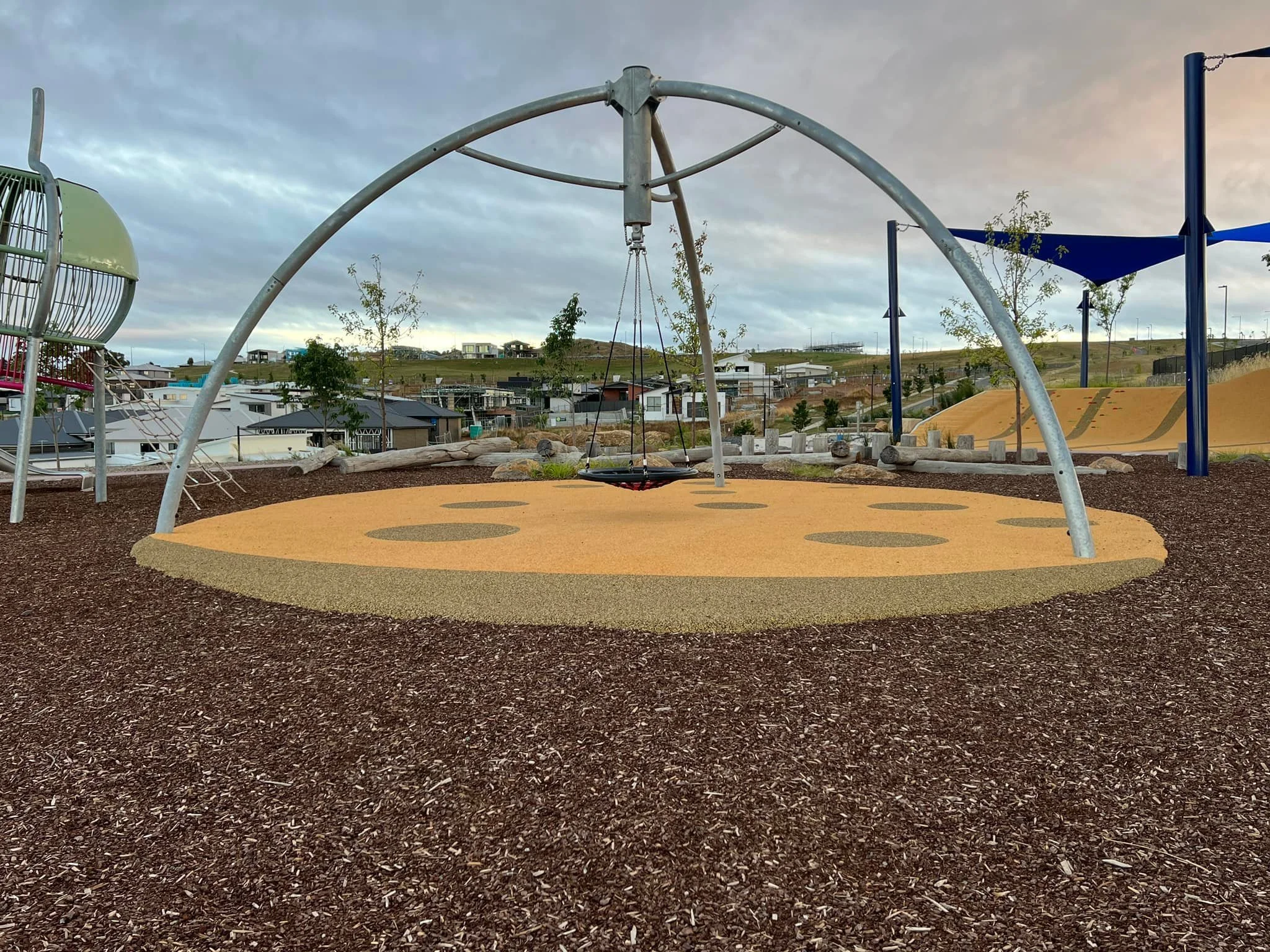 Empty playground with a swing set, slide, and climbing structures on a rubberized surface with houses and trees in the background under a cloudy sky.