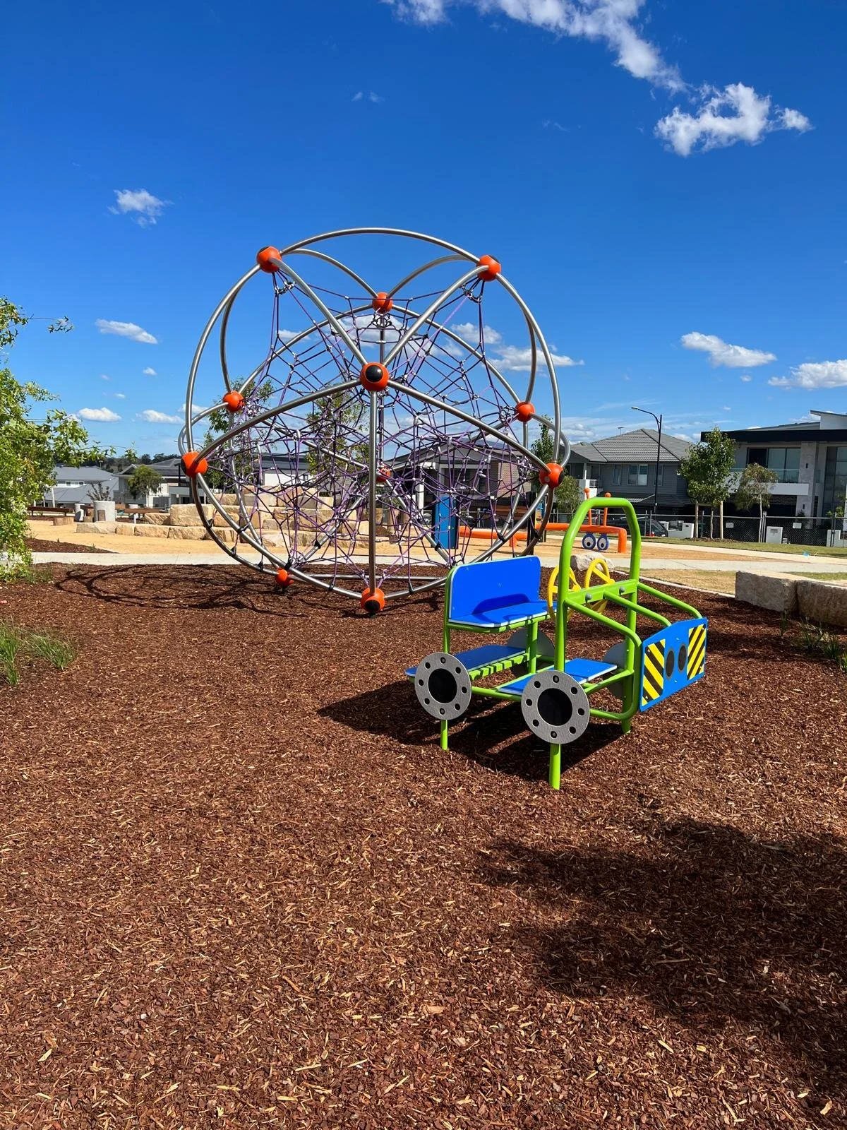Children's playground with a geometric climbing structure and a car-shaped play equipment on a mulch surface, under a blue sky.