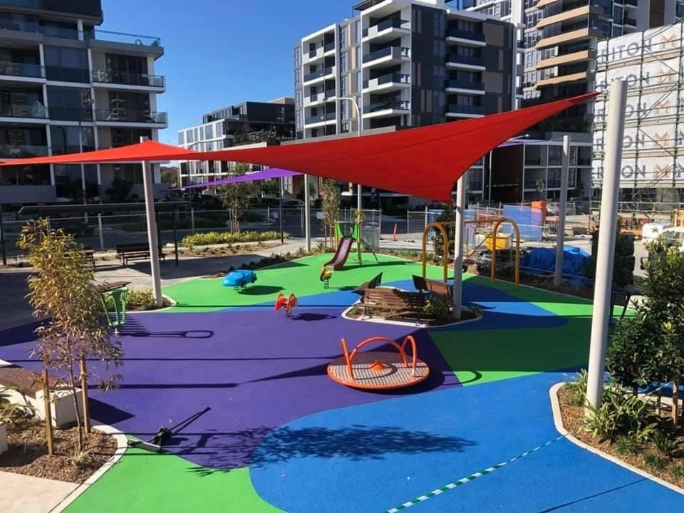 Colorful playground with shade sails, slides, and swings, surrounded by modern apartment buildings.