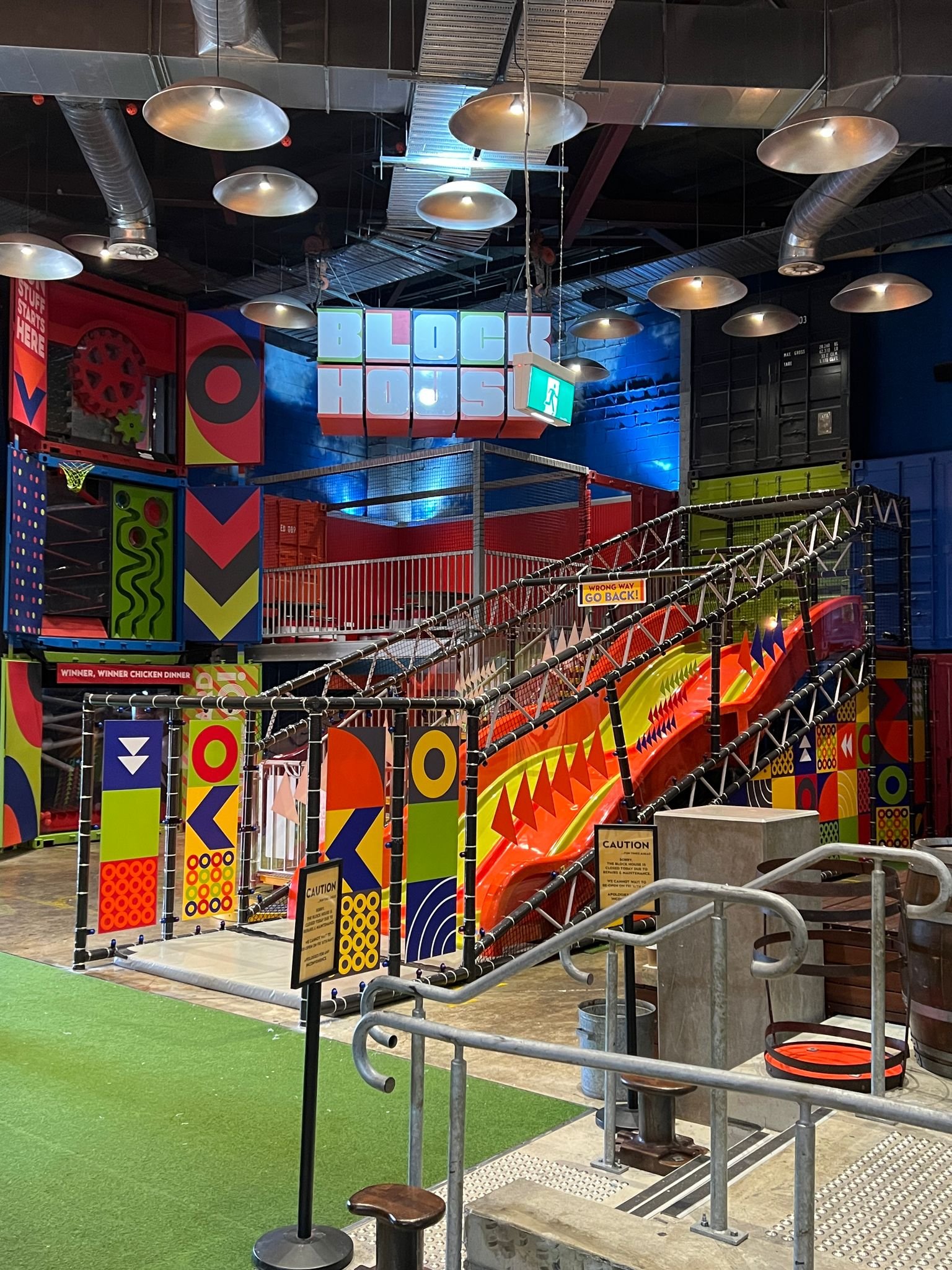 Indoor playground with a colorful slide and climbing structure, featuring a sign that says 'BLOCK HOUSE', safety barriers, and caution signs.