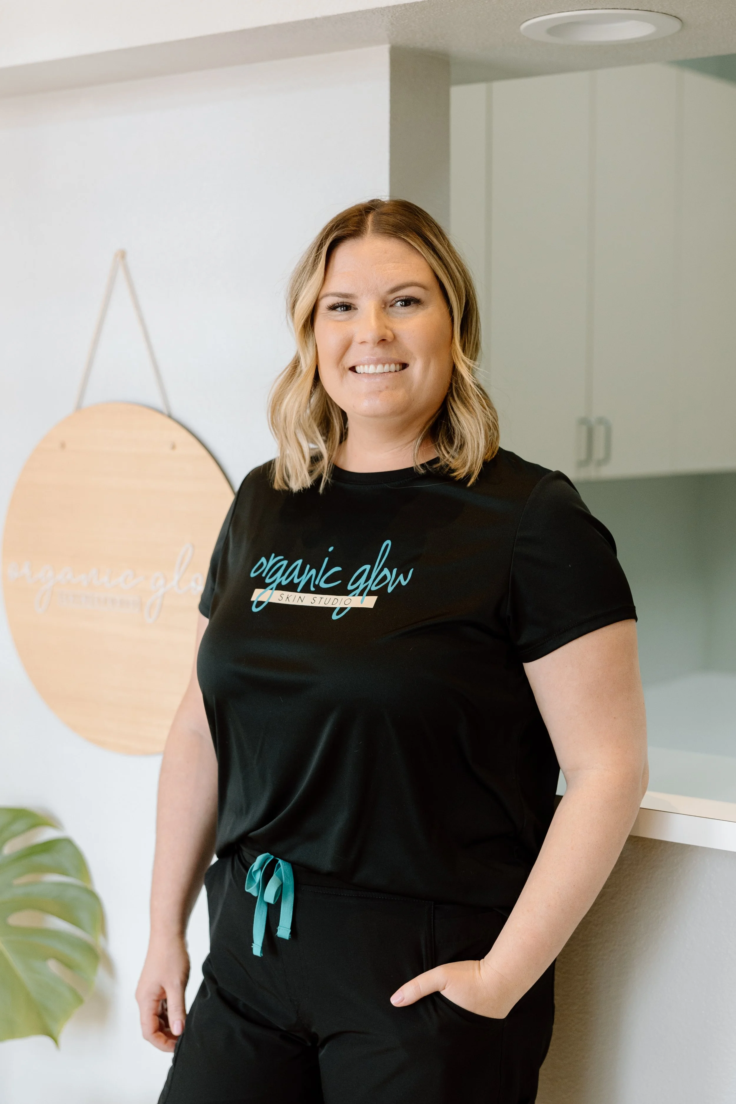 A smiling woman with shoulder-length blonde hair, wearing a black t-shirt with 'organic glow skin studio' written on it, standing indoors in front of a white wall with a wooden sign and a green plant nearby.
