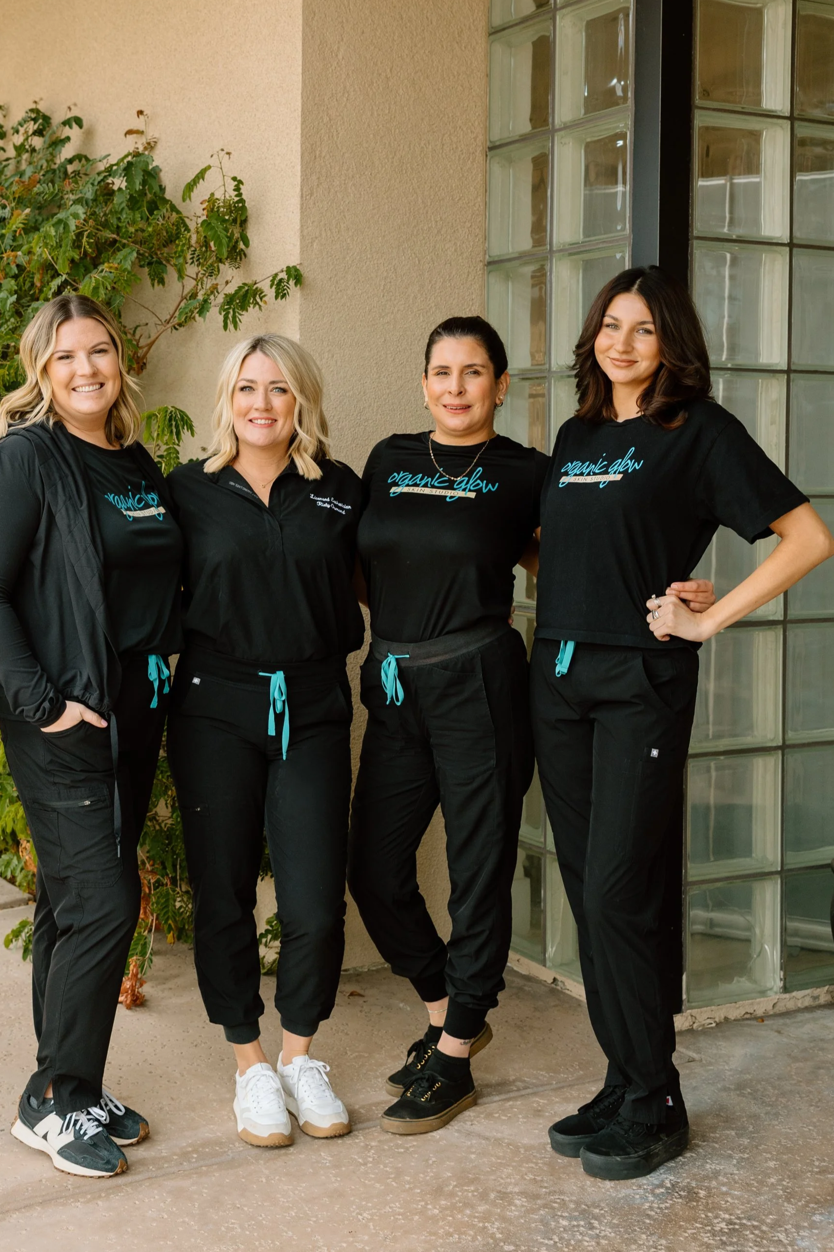 Four women standing together outdoors, wearing black scrubs with the words 'organic glow' on their shirts. They are smiling and standing in front of a beige wall with a glass block window and some greenery.