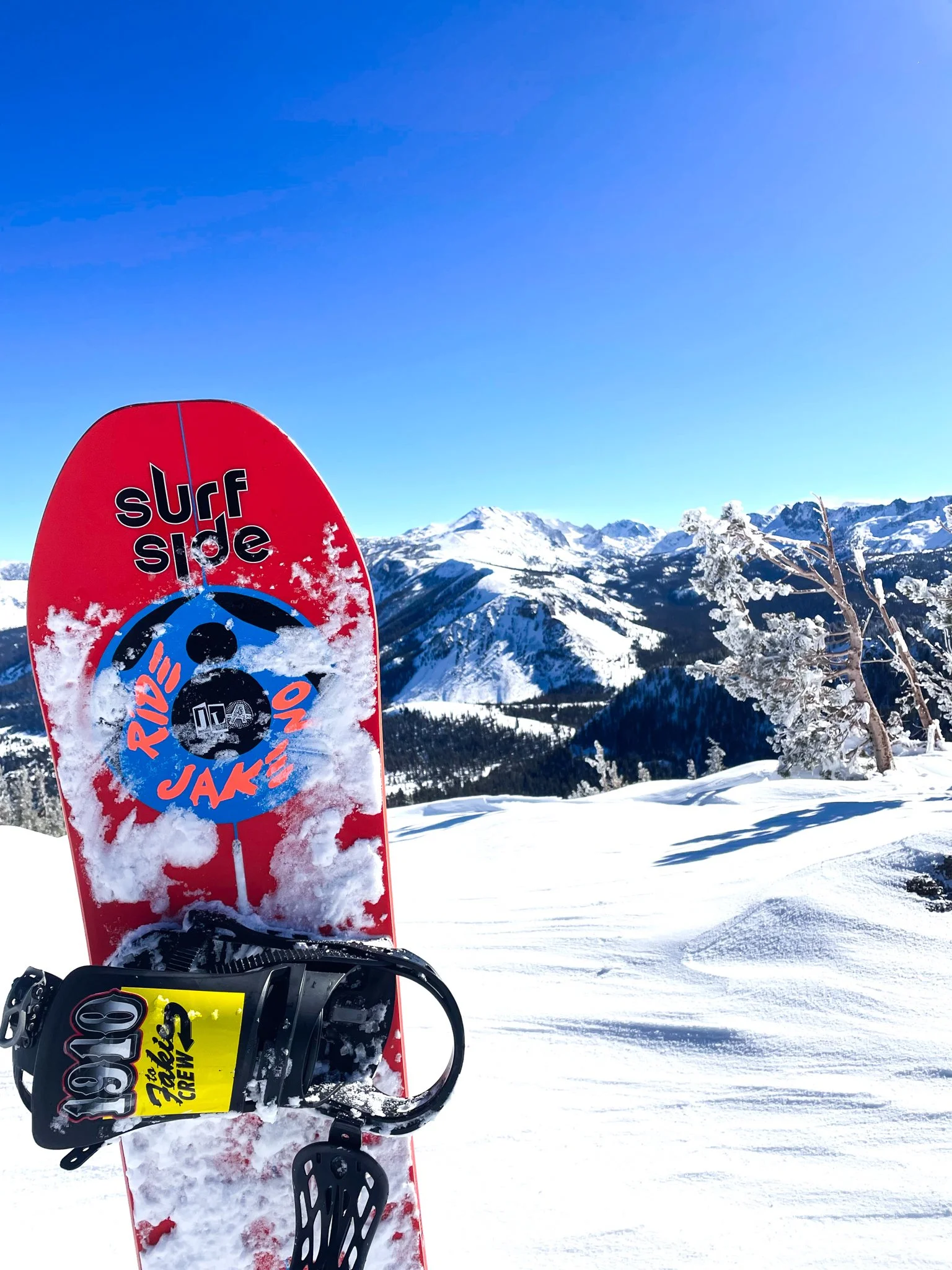 A red snowboard with bindings and snow on it stands upright in the snow, with snow-covered mountains and trees under a clear blue sky in the background.