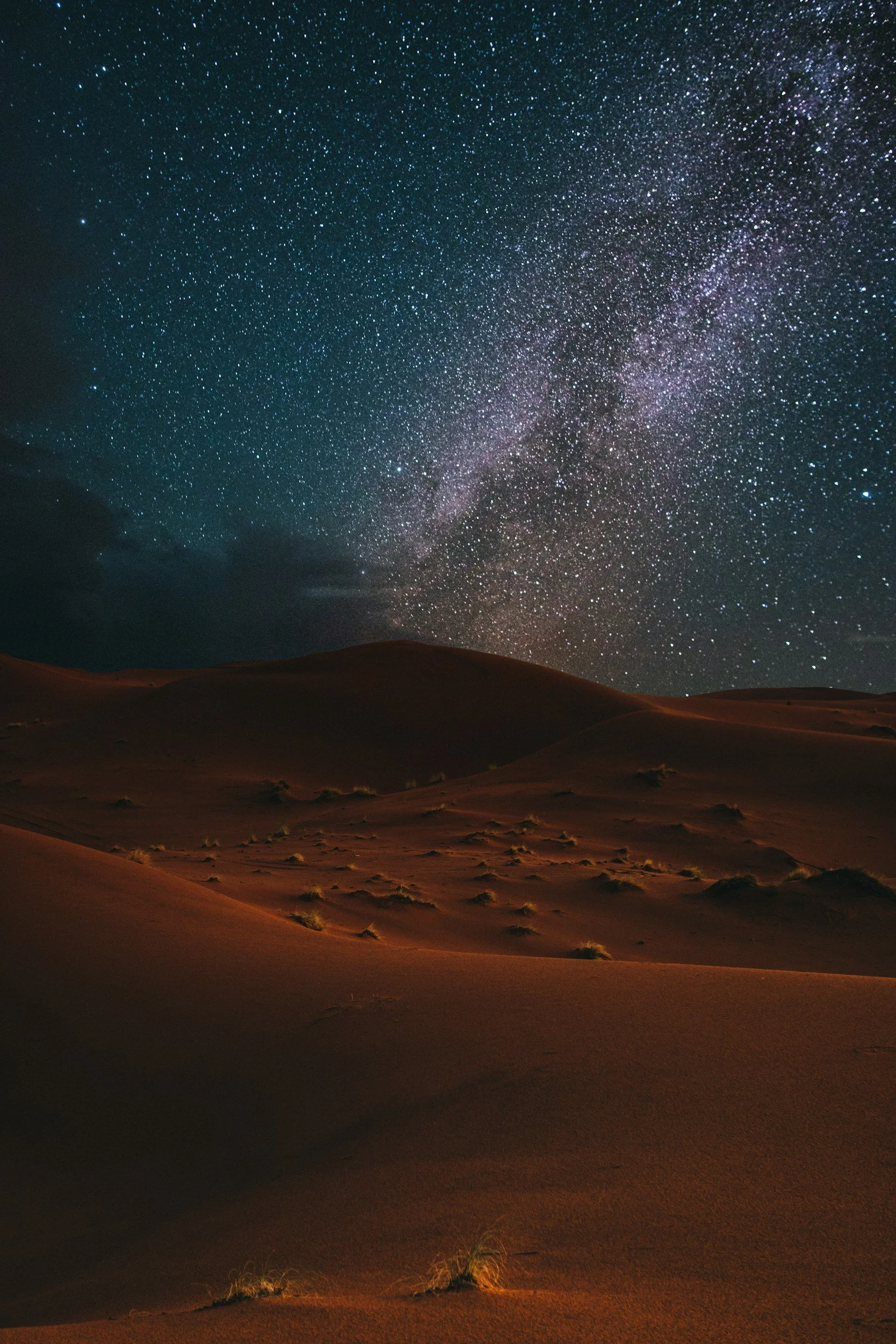 Nighttime desert landscape with sand dunes under a starry sky, including the Milky Way galaxy.