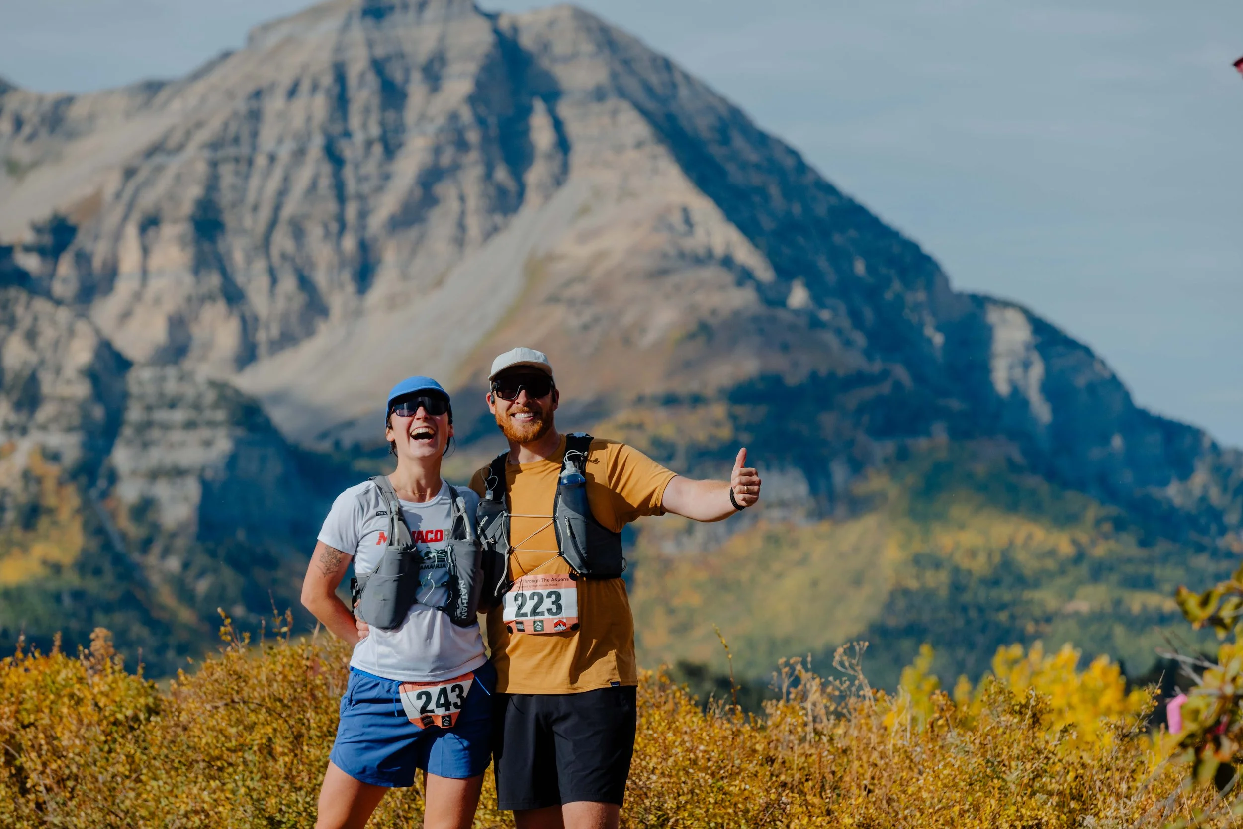 Two smiling trail runners in athletic gear posing in front of a mountain during autumn, with bib numbers 243 and 223.