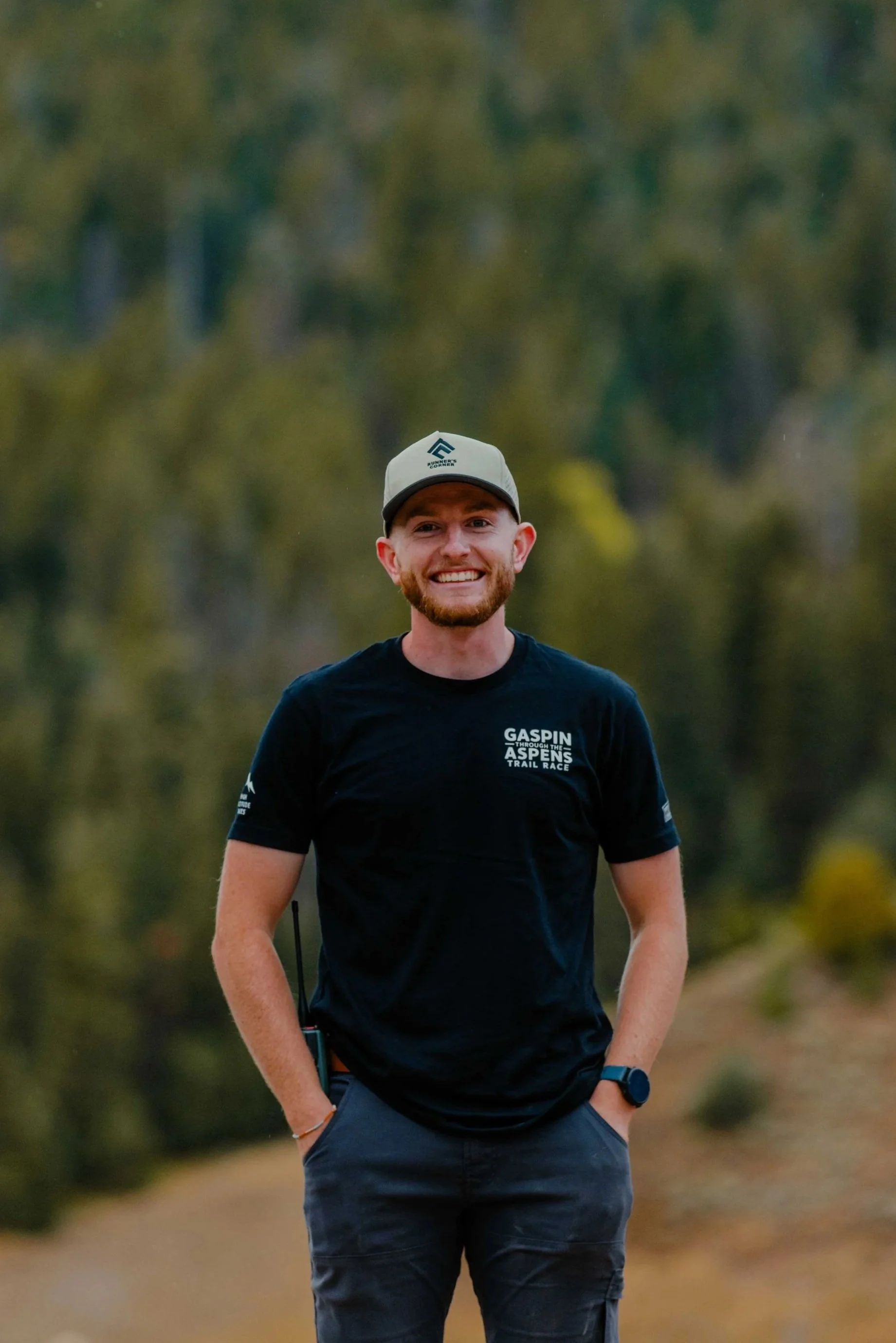 A smiling man standing outdoors in a forested area with trees in the background. He is wearing a black t-shirt, a cap, and a watch.