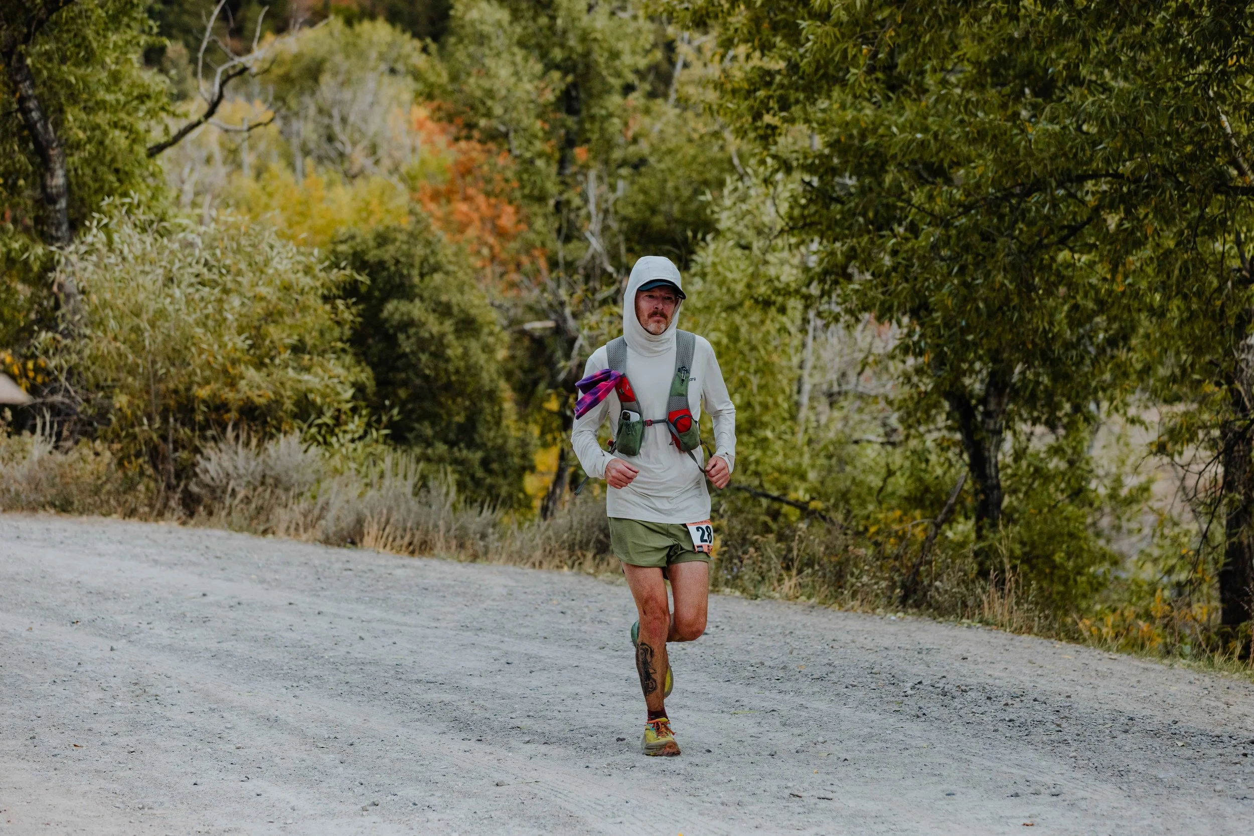 Man running on a gravel trail in a wooded area during autumn, wearing a white hooded long-sleeve shirt, green shorts, a gray hat, and a hydration vest.