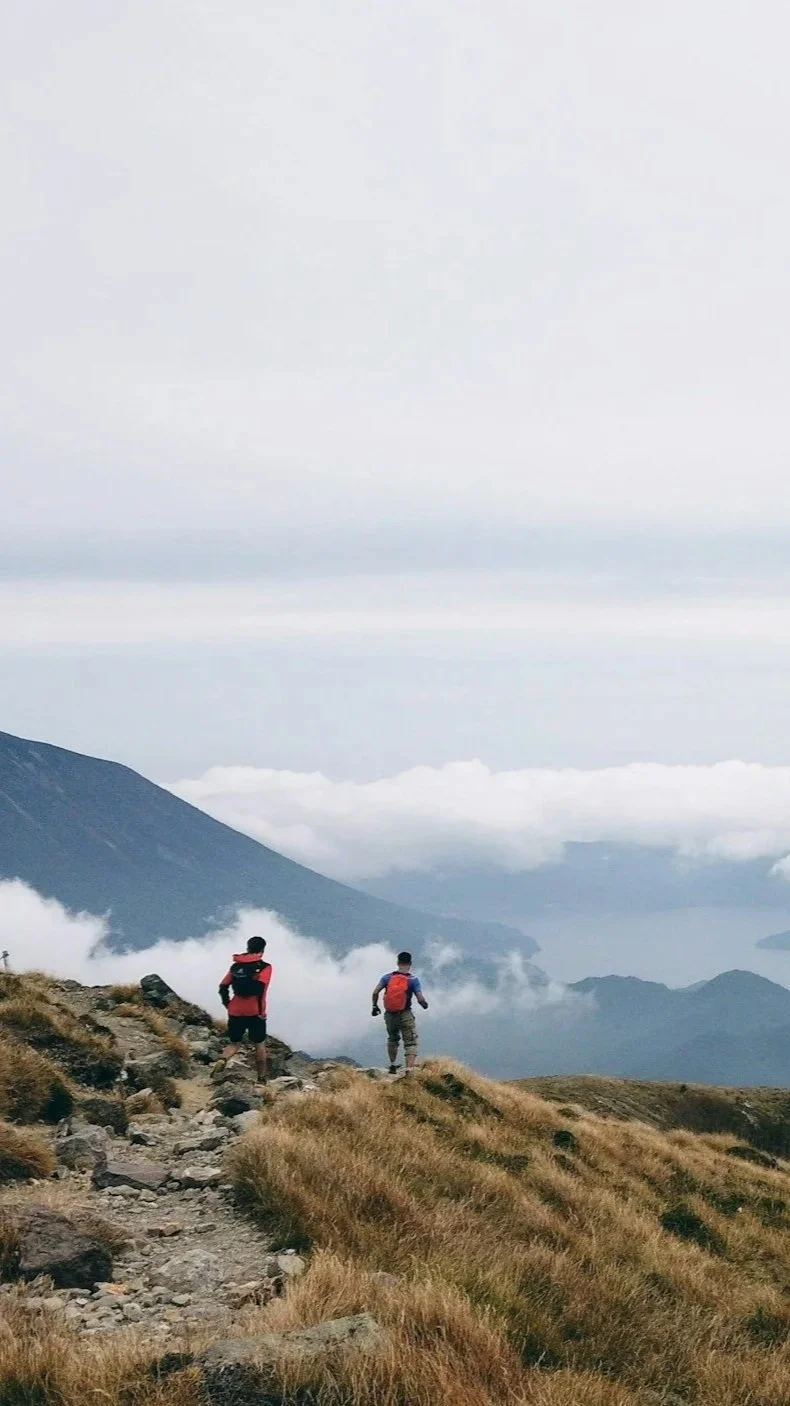 Two hikers with backpacks walking on a rocky trail in a mountainous landscape with clouds and a mountain peak in the background.