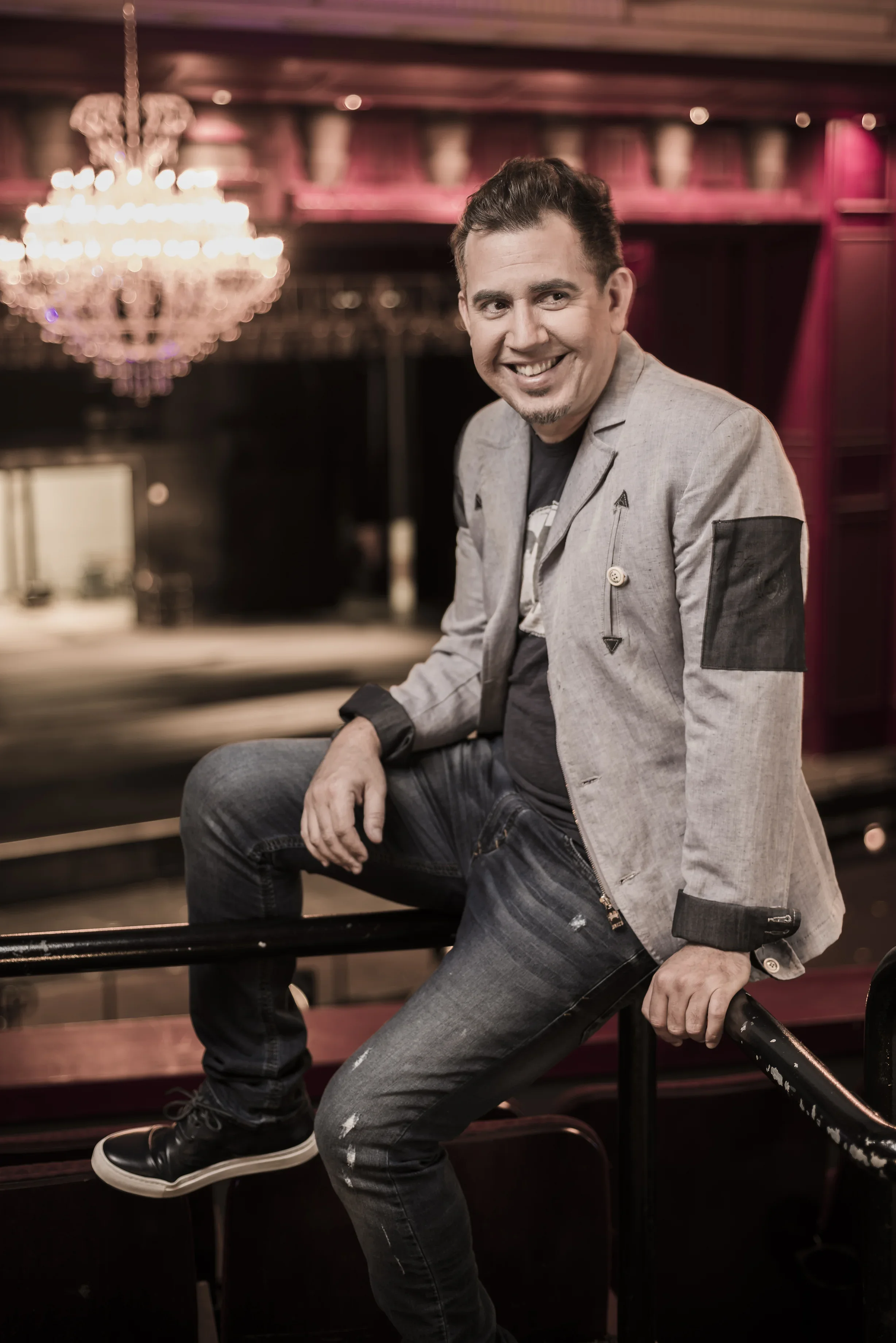 Hombre con chaqueta gris, camiseta negra y jeans oscuros sentado en barandal, sonriendo en un entorno elegante y con candelabro grande en el fondo.