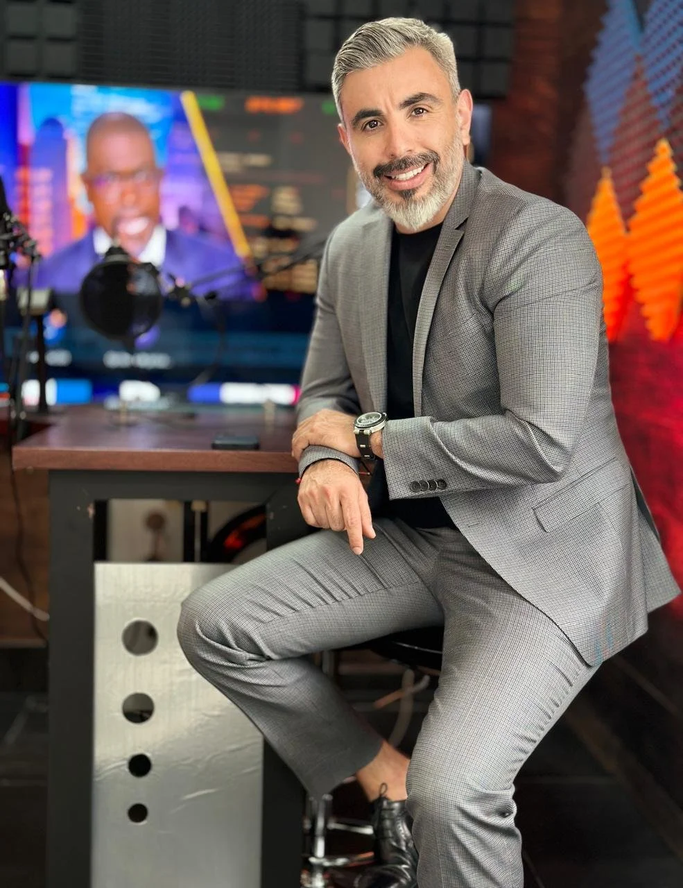 Hombre con barba y cabello gris, vestido con saco gris y pantalón a cuadros, sentado en un escritorio en un estudio de transmisión, con una pantalla grande en el fondo que muestra a otra persona hablando.