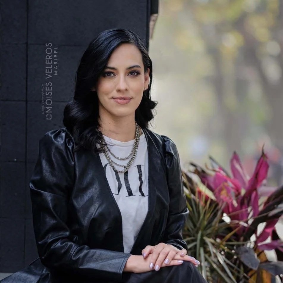 Mujer con cabello oscuro y ondulado, vestida con chaqueta de cuero negra y camiseta blanca, sentada frente a plantas con hojas rosas y verdes, en un fondo borroso con luz natural.