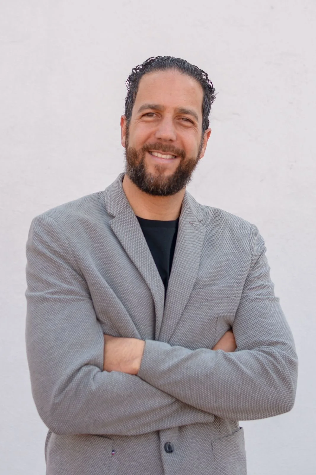 Hombre con barba y cabello rizado, sonriendo, vestido con saco gris y camiseta negra, de pie con brazos cruzados frente a una pared blanca.