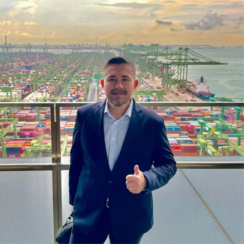 Hombre con traje de negocios de pie en un balcón con vista a un puerto lleno de contenedores y grúas en un atardecer nublado.