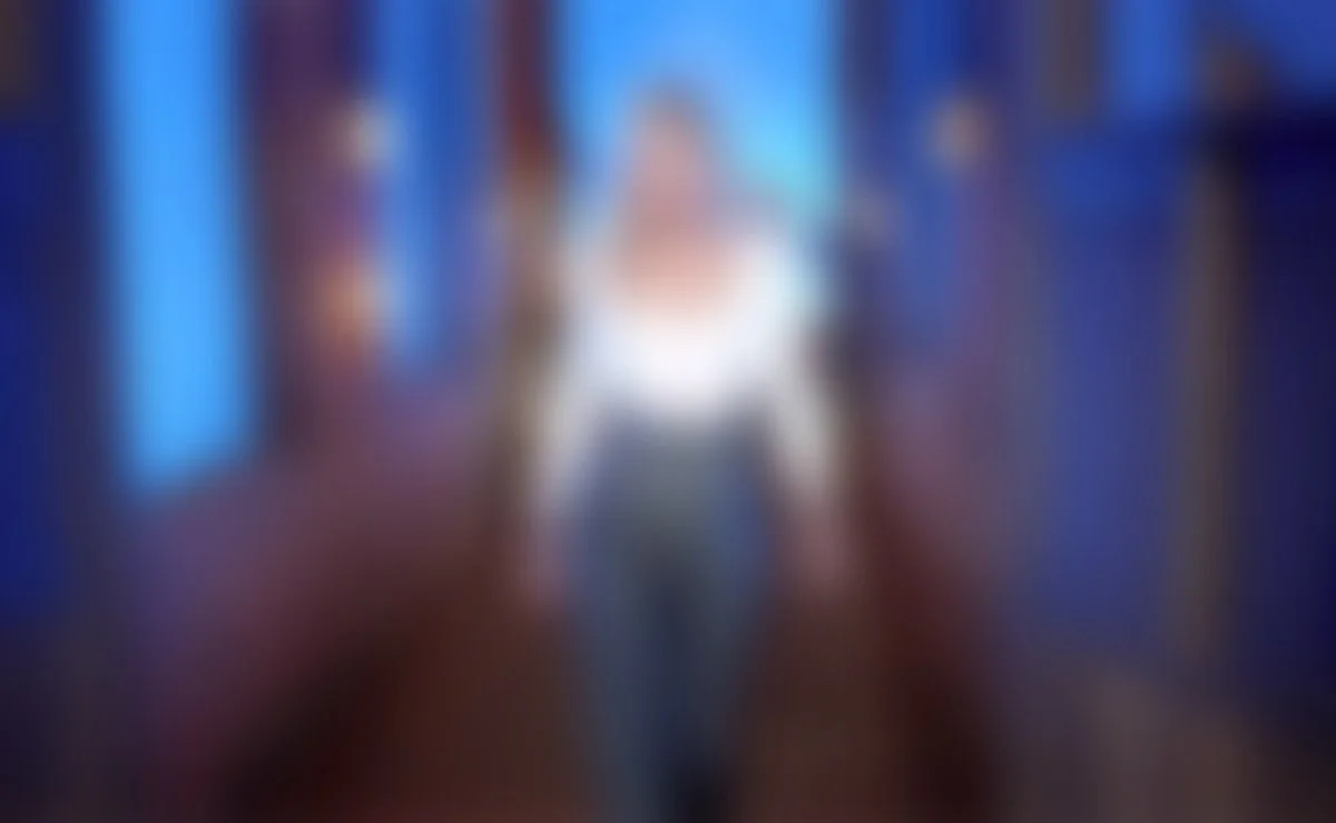 Mujer rubia con vestido blanco y pantalones grises camina en un pasillo con paredes de ladrillo y ventanas con contraventanas en un ambiente con iluminación cálida y azul.