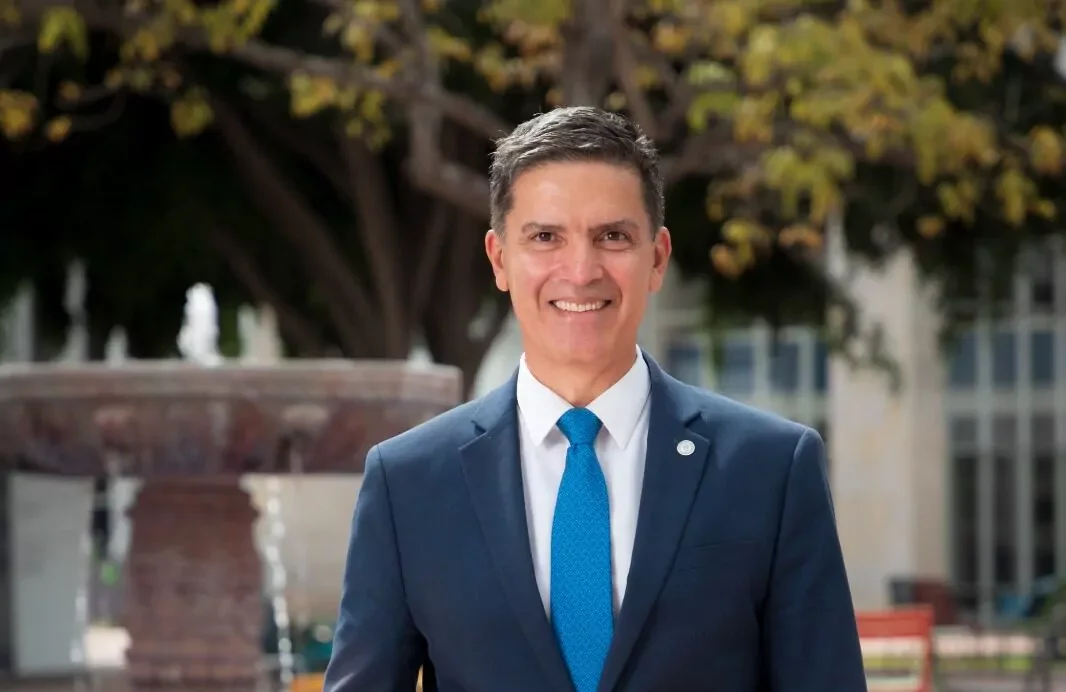 Hombre con traje azul y corbata azul, sonriendo, en un entorno exterior con árbol y fuente.