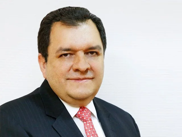 Retrato de un hombre con piel clara, cabello oscuro y peinado hacia atrás, usando un traje negro, camisa blanca y corbata roja, contra un fondo blanco