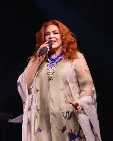 Mujer con cabello rizado y color rojo, disfrazada con vestido beige y un collar azul, sosteniendo un micrófono y sonriendo en un escenario oscuro.