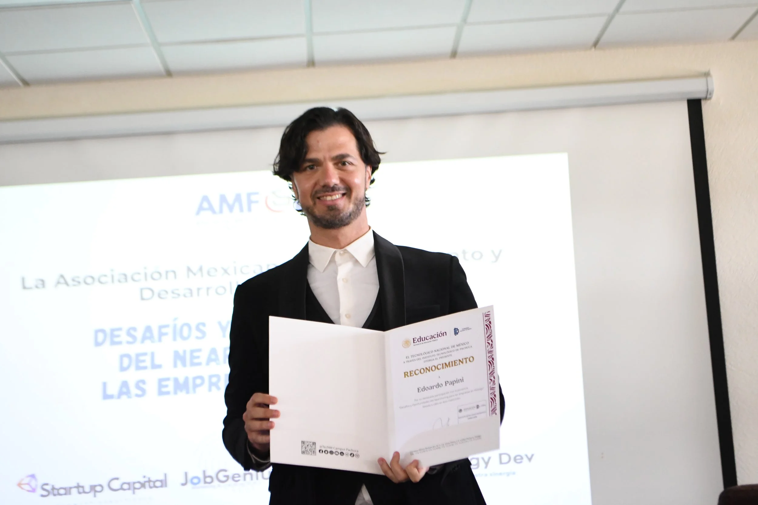 Hombre en atuendo formal sosteniendo un certificado de reconocimiento, sonriendo en un evento de premiación.