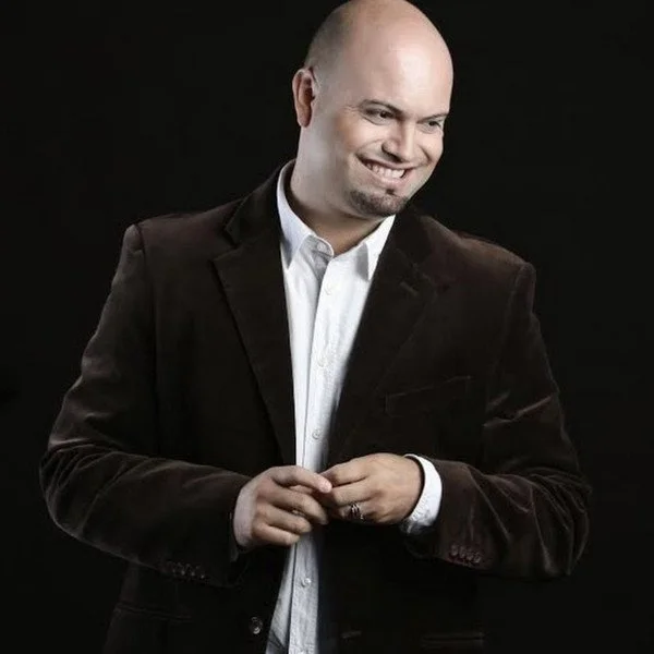 Hombre sonriendo, vestido con chaqueta oscura y camisa blanca, en un fondo negro.