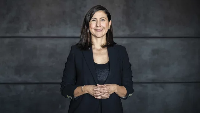 Mujer sonriendo, vestida con traje negro y blusa, de cabello castaño oscuro, en un fondo gris