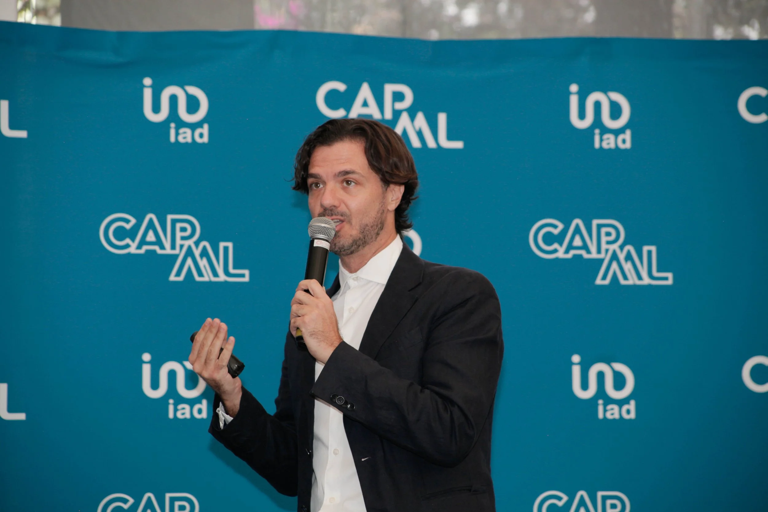 Hombre con barba y cabello castaño oscuro, usando saco negro y camisa blanca, hablando en micrófono en un evento con fondo azul que tiene logotipos de CAP AML e IAD.