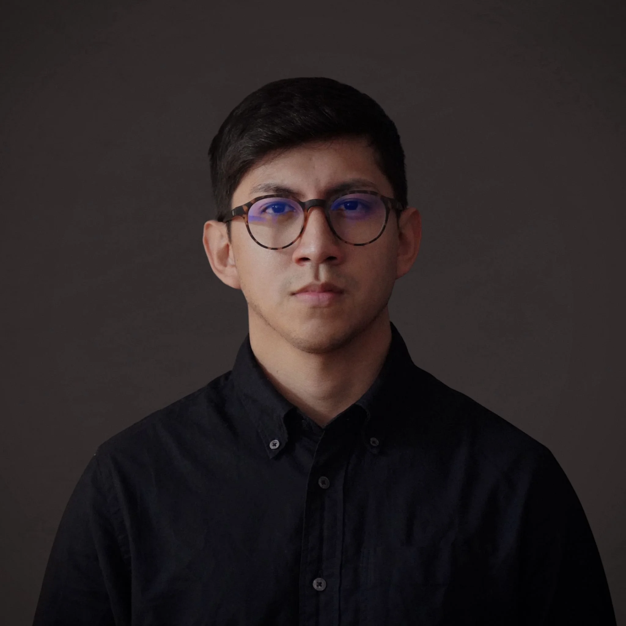 Retrato de un joven con gafas, cabello oscuro y camisa negra, mirando hacia la cámara contra un fondo oscuro.