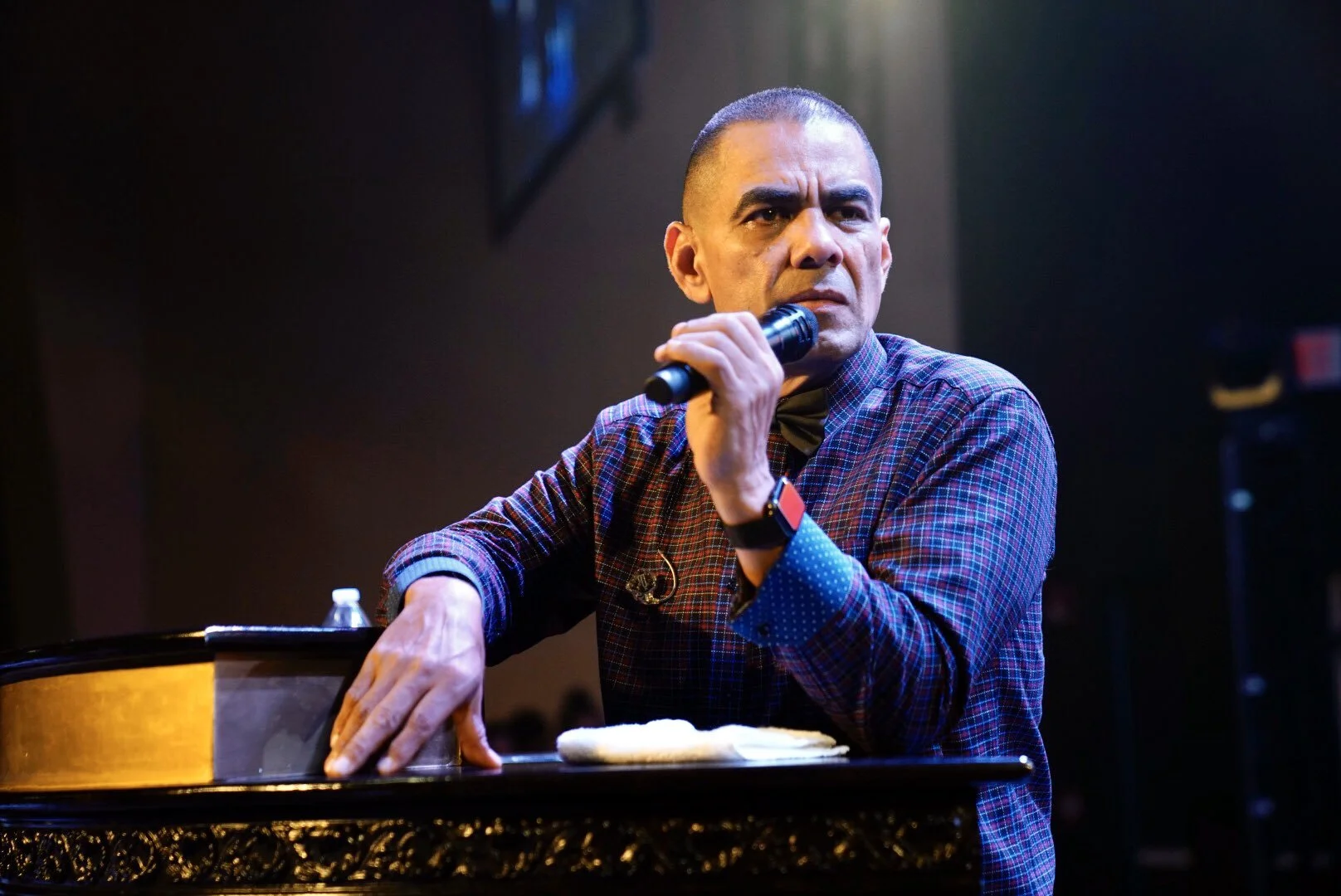 Un hombre con traje claro sosteniendo un micrófono en un concierto, sonriendo y gesticulando con la mano, mientras el público lo aplaude.