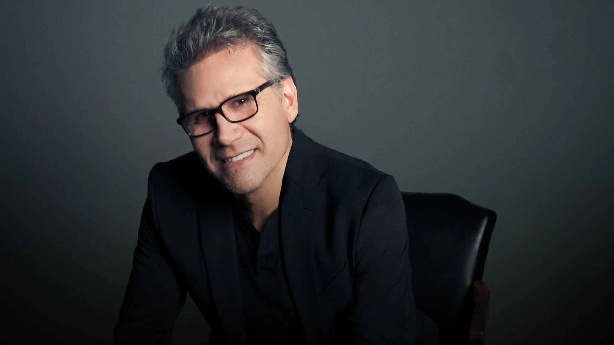 Hombre con gafas y cabello canoso, vestido con saco negro, sentado en una silla negra en un fondo gris oscuro