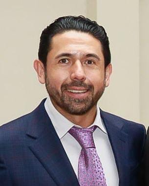 Hombre con traje azul y corbata púrpura, sonriendo, fondo neutro.