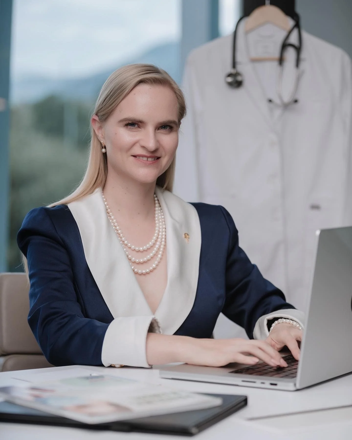 Mujer con pelo rubio y ropa formal trabajando en una computadora en una oficina, con un uniforme de médico y estetoscopio colgado en la pared.