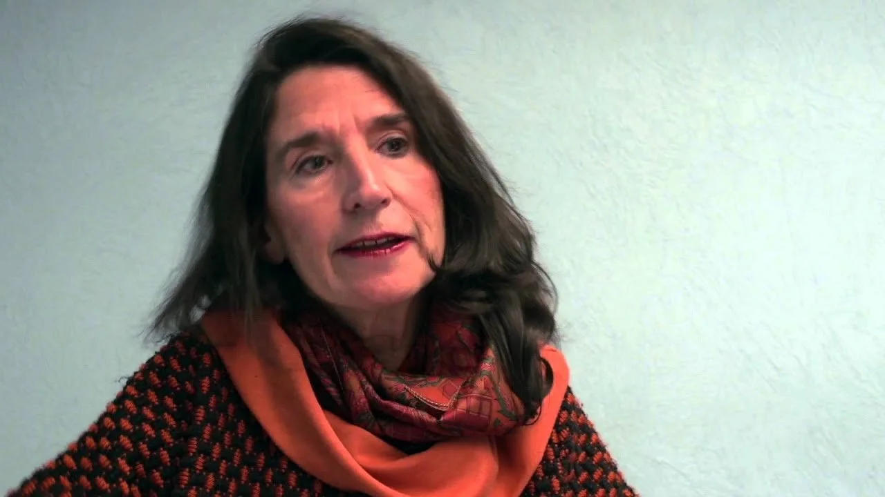 Mujer de cabello castaño oscuro y ojos claros, con expresión de conversación, lleva un suéter negro con puntos rojos y una bufanda anaranjada con detalles, en un fondo de pared blanca.