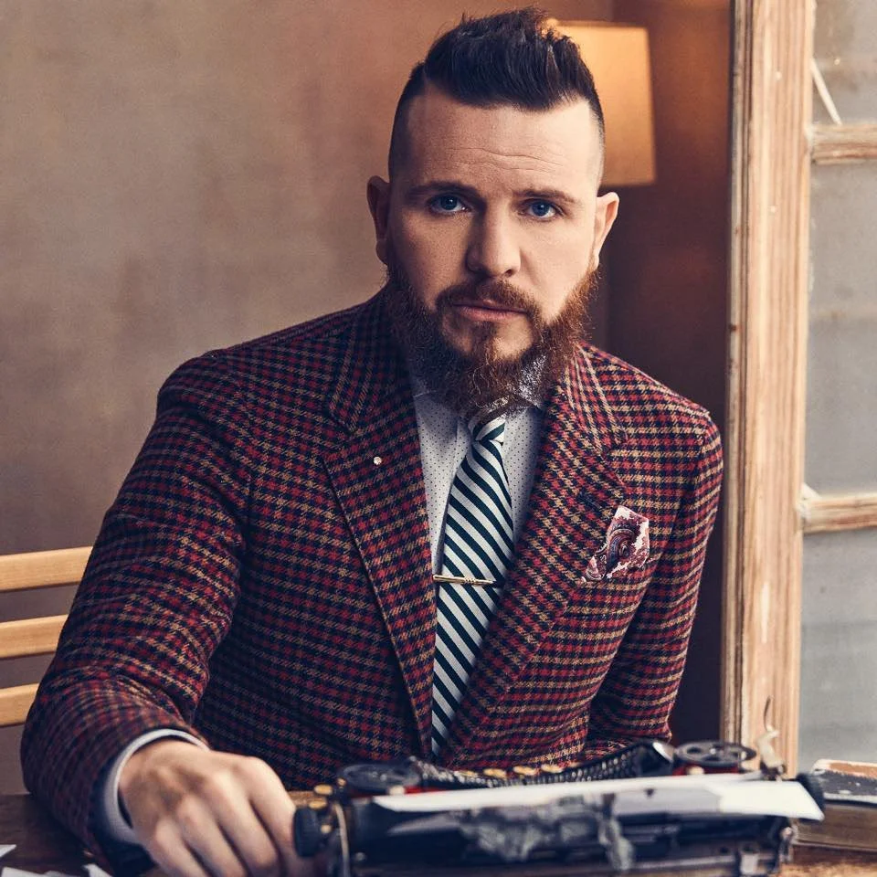 Hombre sonriendo, con barba y un peinado moderno, vestido con saco rojo a cuadros y camisa blanca, posando en un fondo gris oscuro.