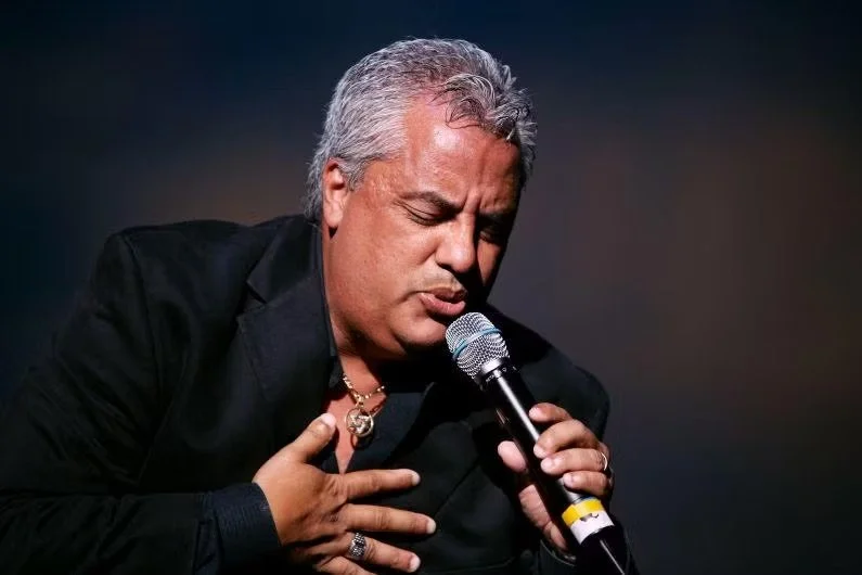 Hombre con cabello gris, vestido con traje negro, cantando o hablando con un micrófono en mano, con expresión emocional.