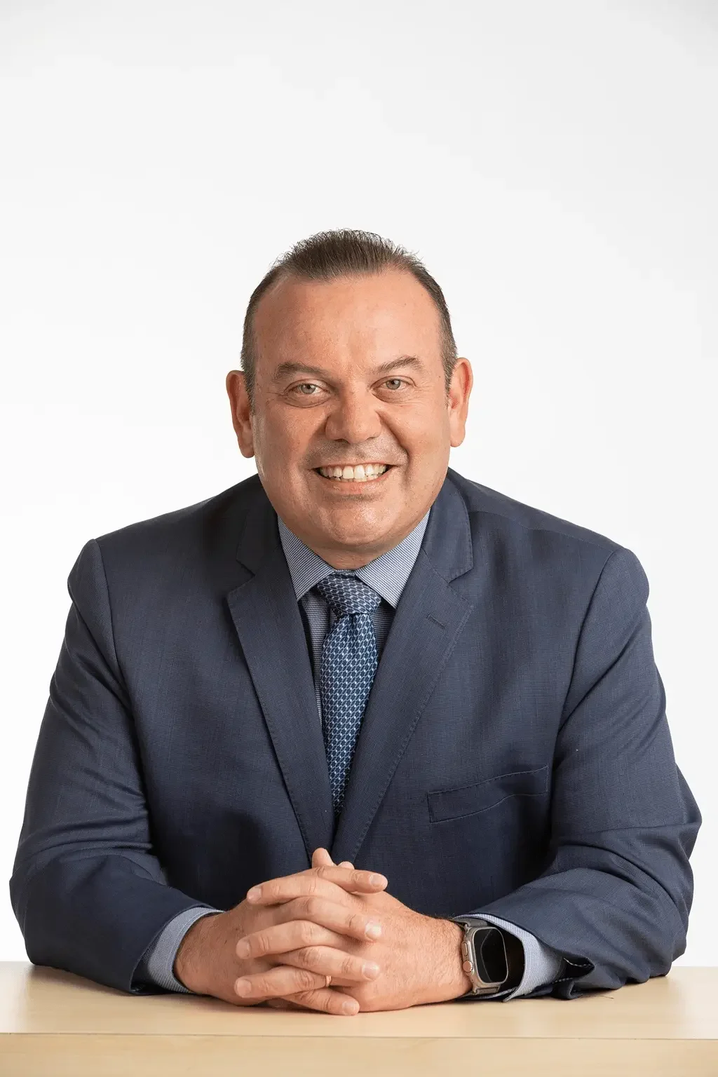 Hombre de negocios sonriendo, vestido con traje y corbata, sentado en una mesa con las manos juntas y una expresión amistosa.